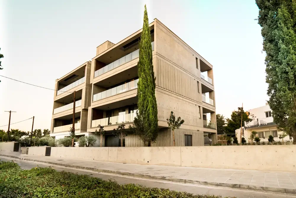 Modern multi-story apartment building with balconies, concrete exterior, surrounded by trees and a low wall, in a suburban neighborhood.