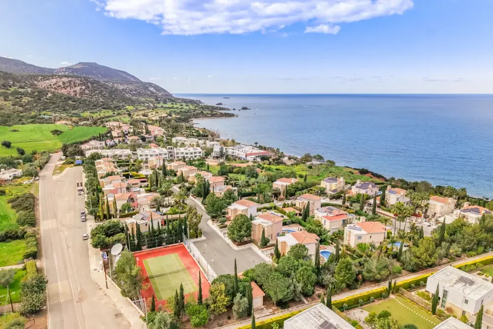 A coastal residential neighborhood with houses, a tennis court, and an ocean view, surrounded by greenery and mountainous terrain in the background.