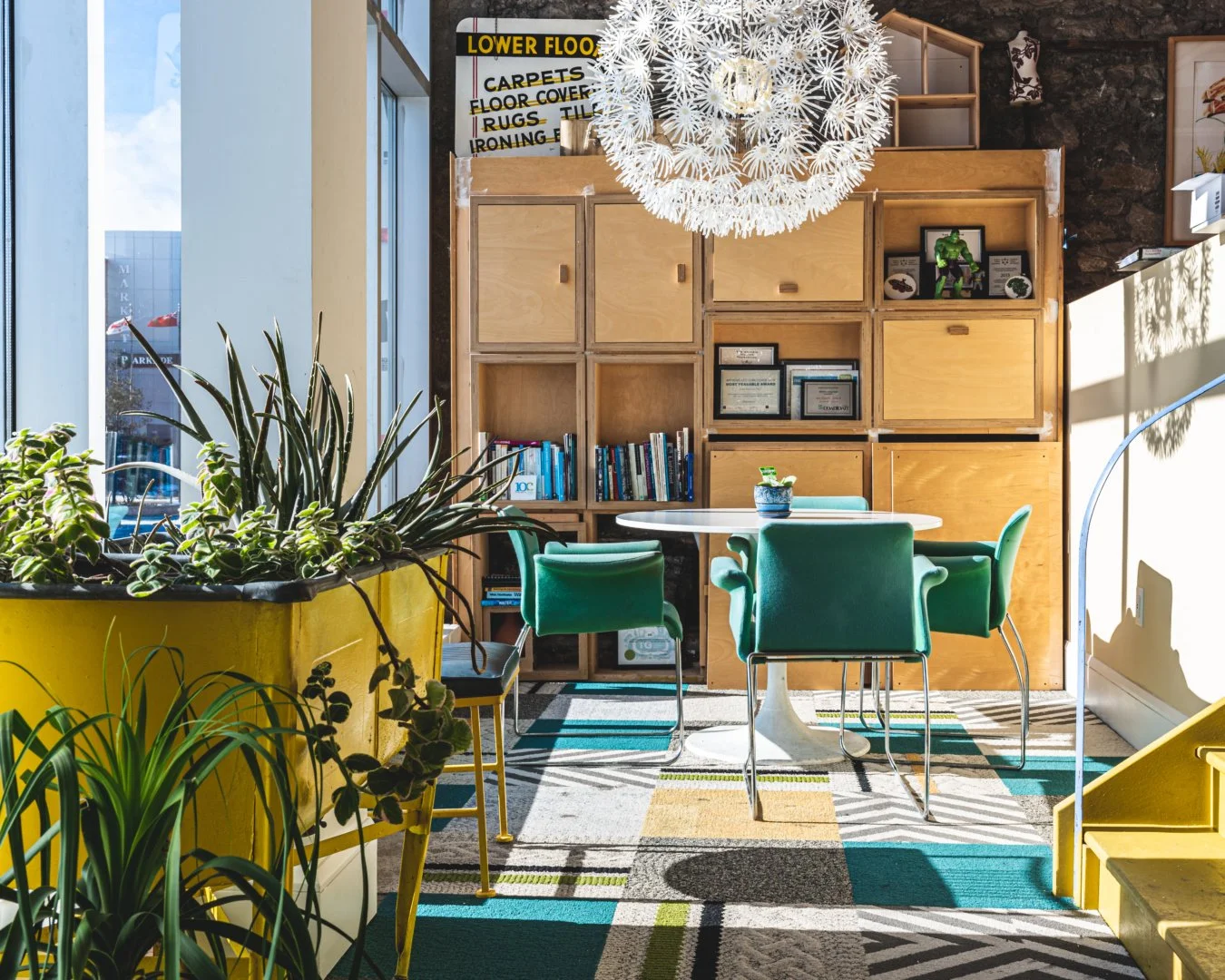 A room with large windows letting in sunlight, a round white table surrounded by five green velvet chairs, green plants in yellow planters, a wooden bookshelf, and a large decorative white spherical chandelier.