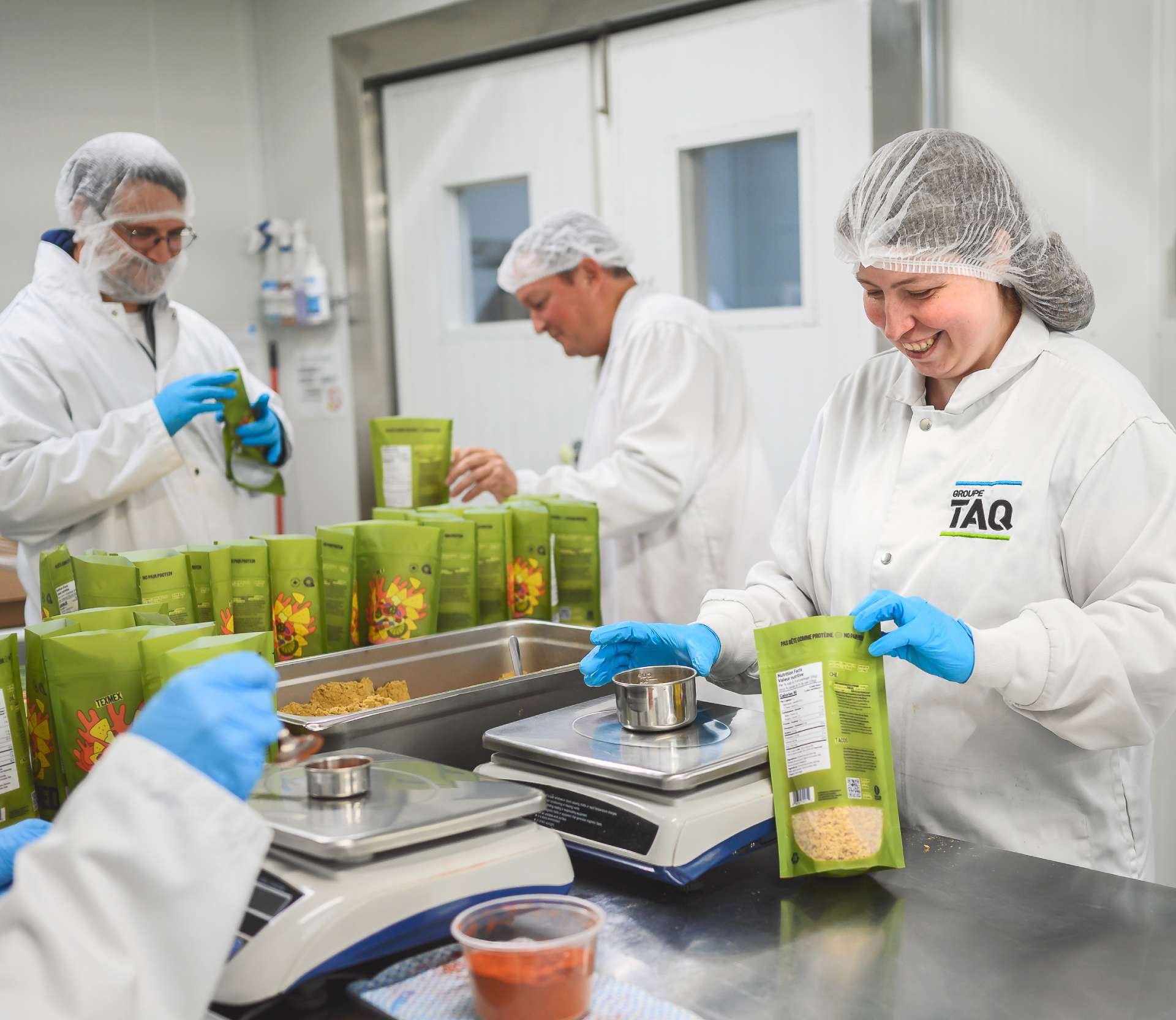 People working in a food production or packaging facility, wearing white coats, hairnets, and face masks, packaging food products in green bags, with digital scales on the table.