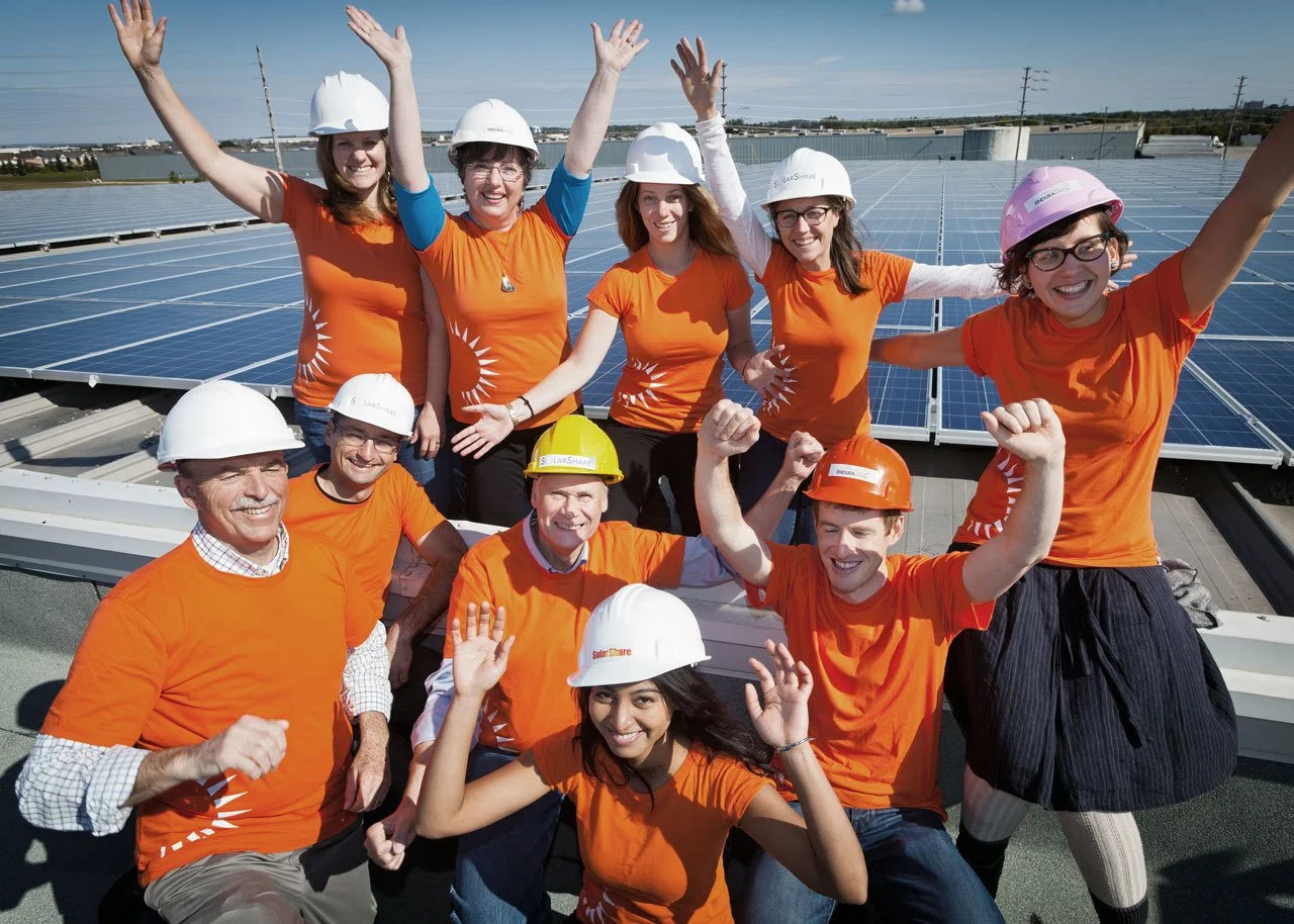 Group of nine people wearing orange shirts and safety helmets celebrating on a rooftop with solar panels.