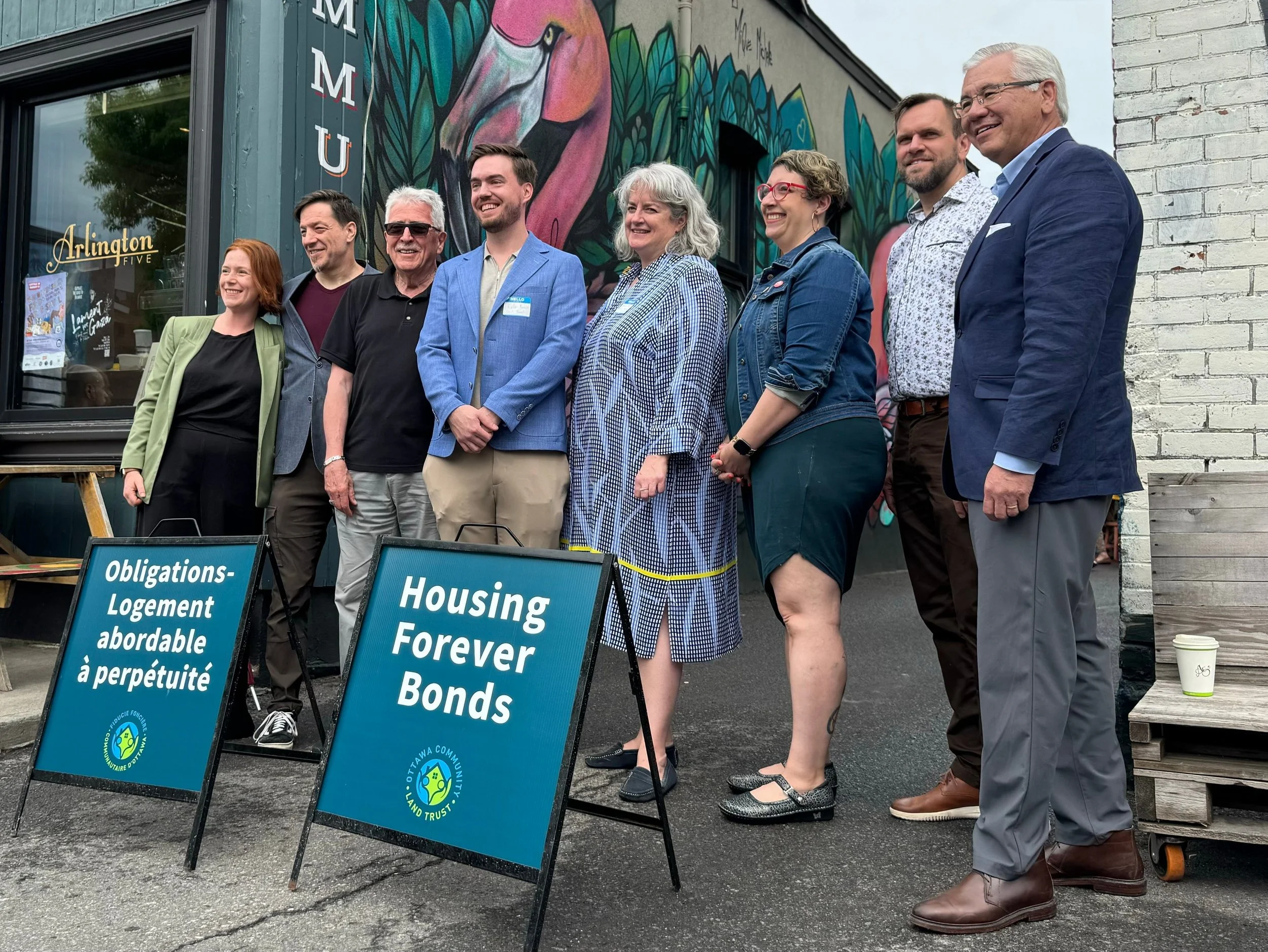 Group of nine people standing outside in front of a colorful mural, smiling. Two signs read 'Obligations-Logement abordable à perpétuité' and 'Housing Forever Bonds.' The individuals are dressed in casual and business attire, and there is a coffee cup on the ground.