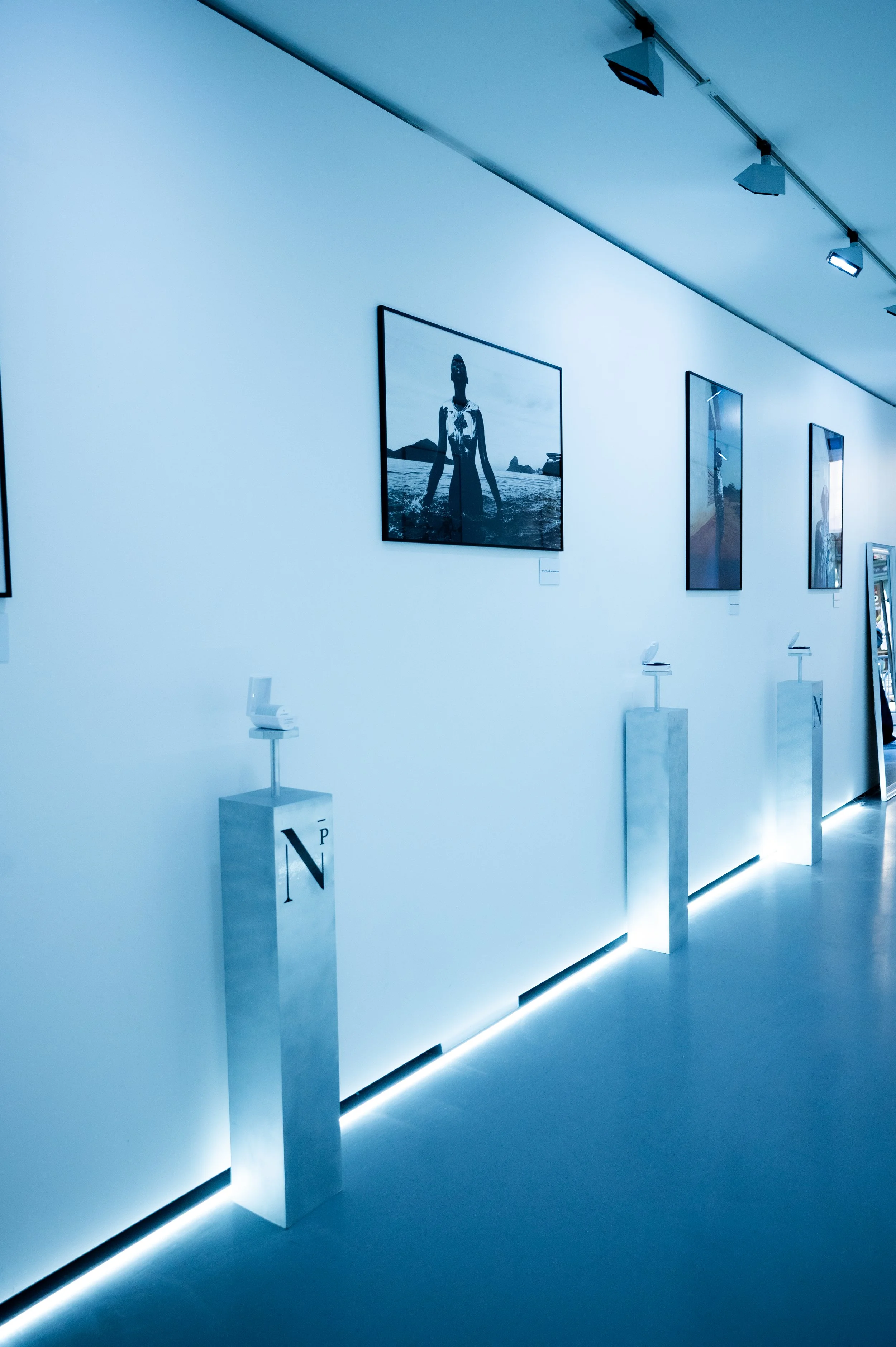 Une galerie d'art avec des peintures encadrées suspendues à un mur blanc, éclairée par des projecteurs au plafond et des éclairages indirects au sol.