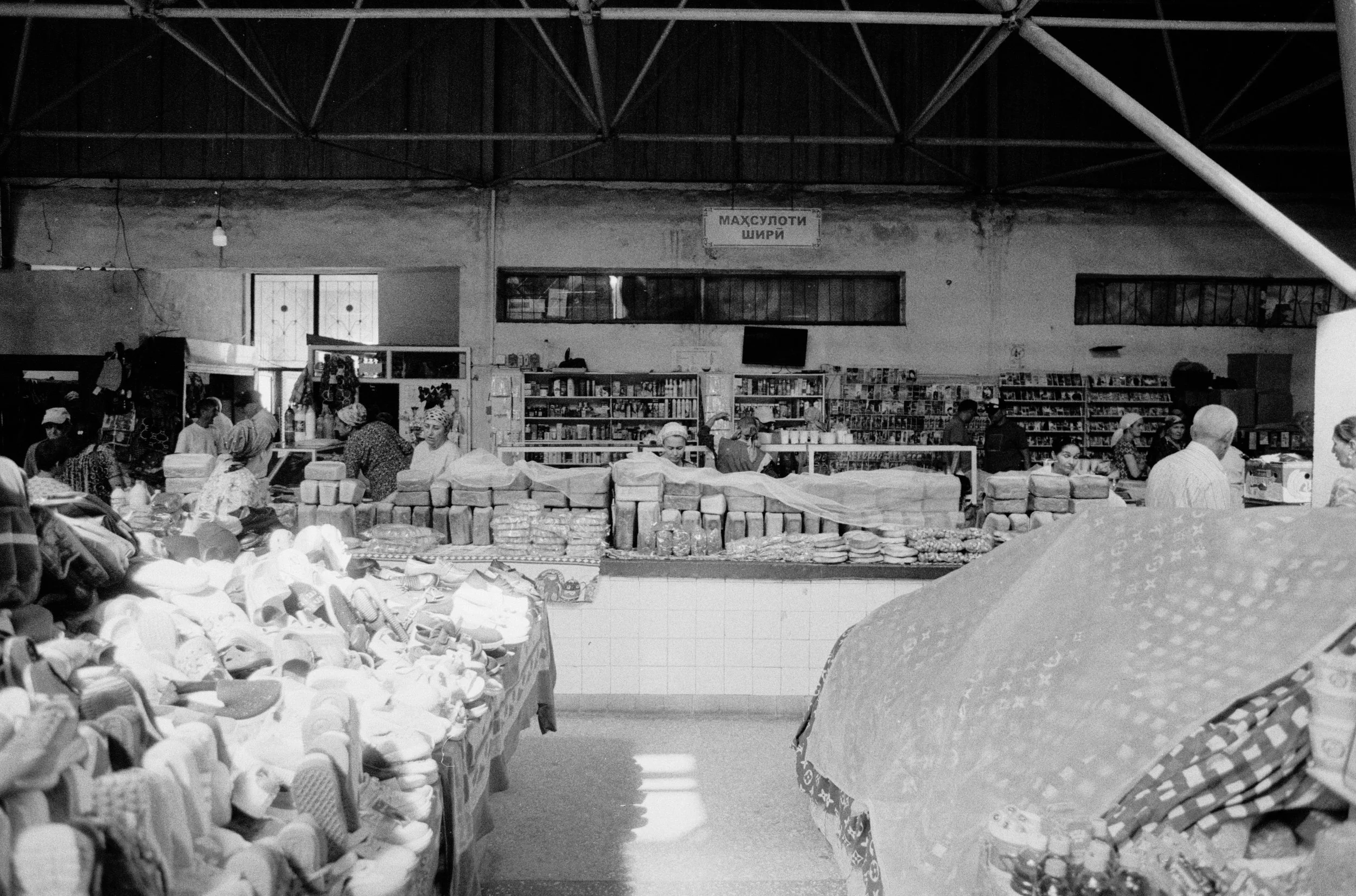 Marché intérieur avec des vendeurs et des clients, étals de produits comme chaussures et autres marchandises, dans un bâtiment ancien. Sign visible avec écriture en alphabet cyrillique.