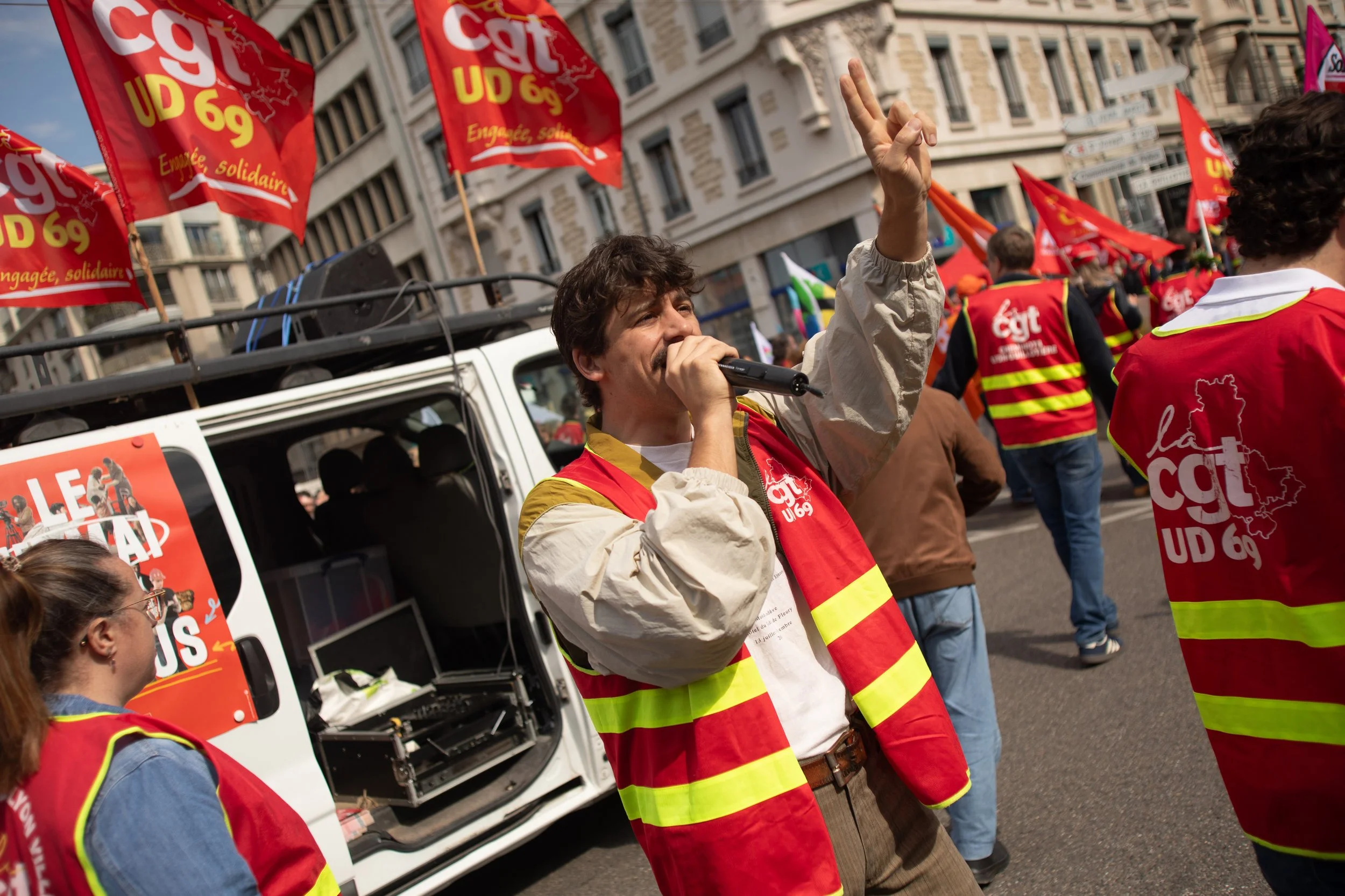 Manifestants lors d'une protestation, portant des gilets de sécurité rouges et jaunes avec le logo de la CGT, entourés de drapeaux rouges avec le texte 'CGT UD 69' et une carte de France, dans une ville avec des bâtiments en arrière-plan.