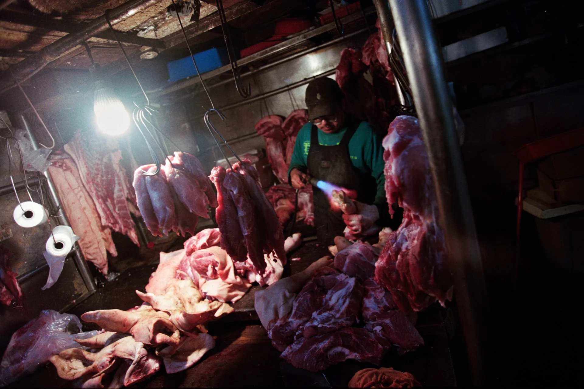 Un boucher travaille dans une boucherie, entouré de carcasses de viande suspendues, sous un éclairage brillant.