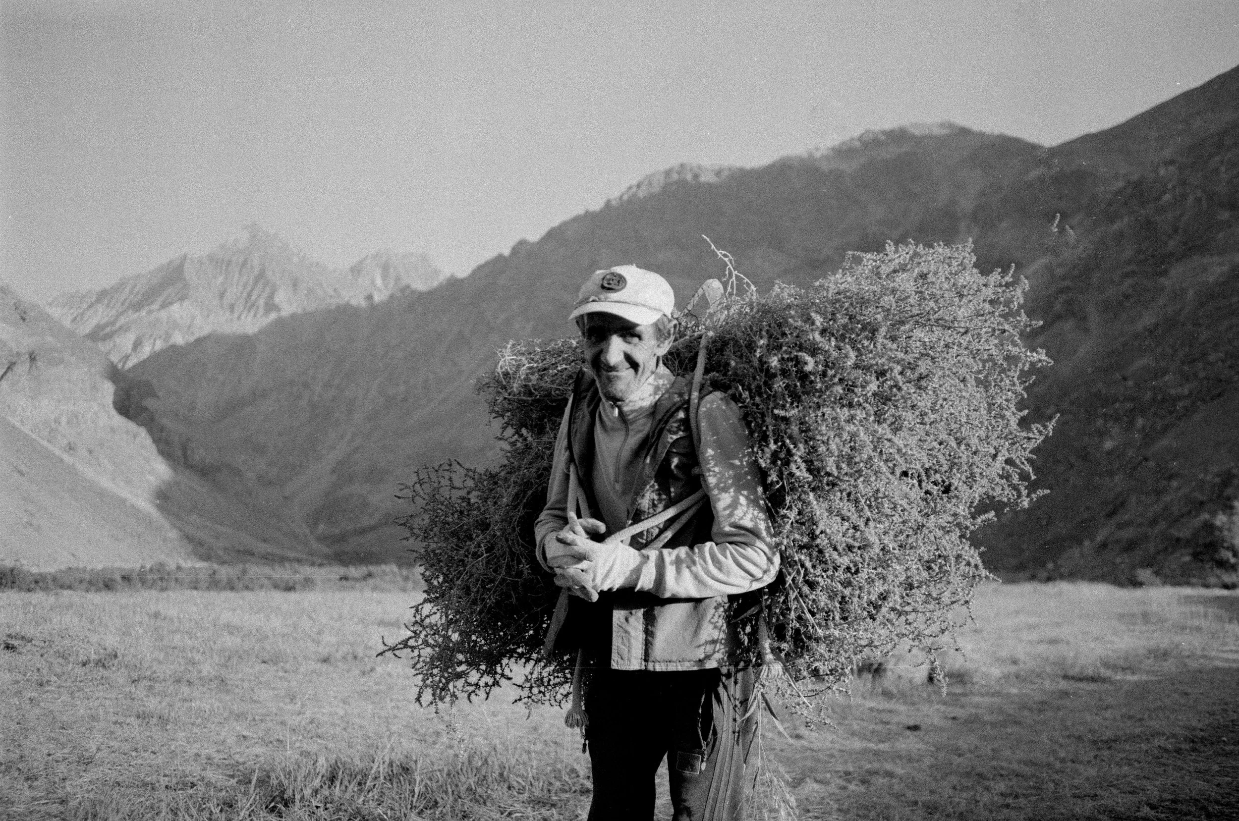 Homme souriant portant un sac à dos, tenant un bouquet de branches ou de feuillage, dans un paysage de montagnes et de champs en arrière-plan.
