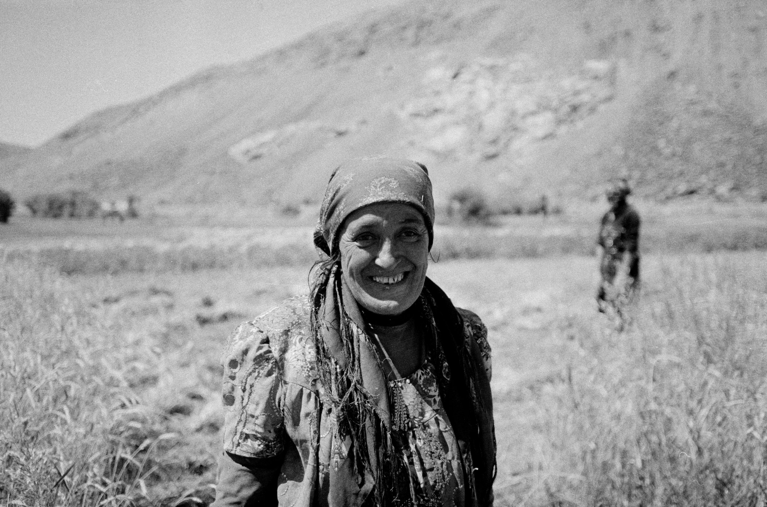 Portrait d'une femme souriante portant un foulard, à l'extérieur dans un paysage montagneux avec herbes hautes, en noir et blanc.