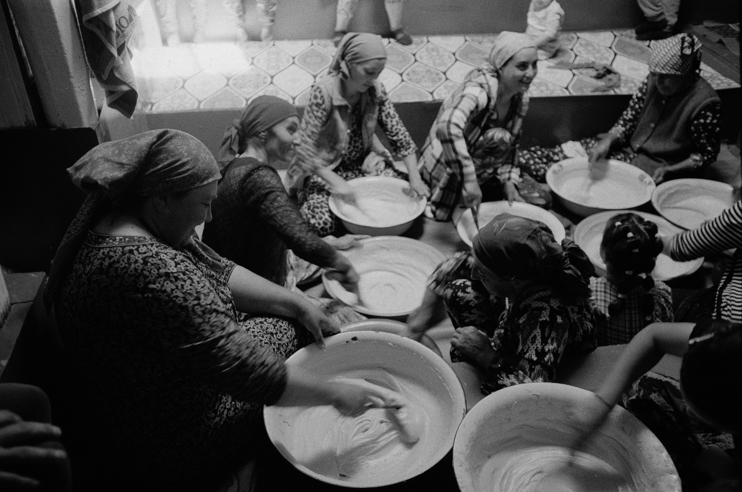 Des femmes portant des foulards colorés, assises en cercle, sont en train de préparer une pâte ou un aliment dans de grands bols, dans une cuisine ou un espace de cuisine.
