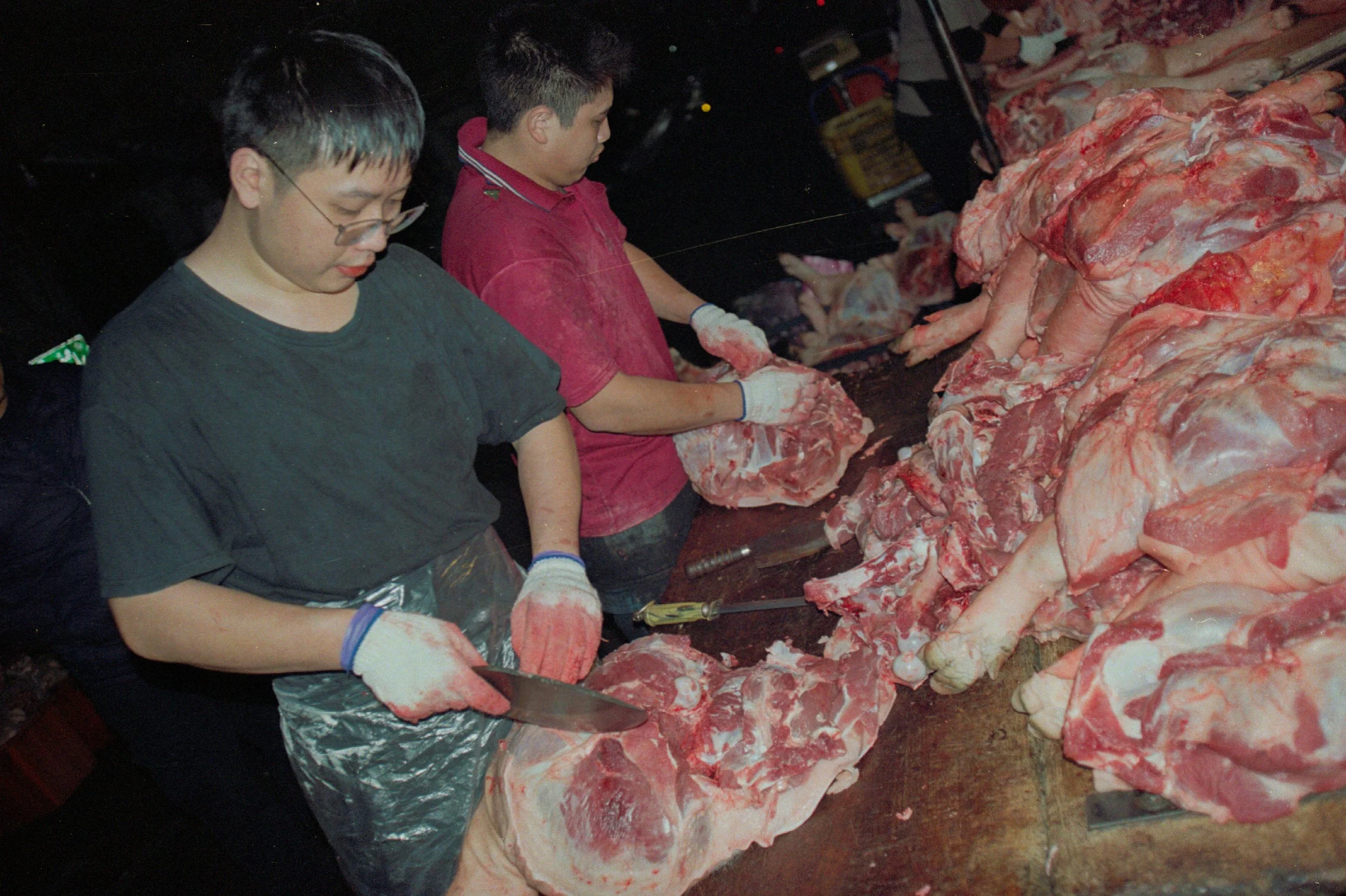 Deux hommes travaillent la viande dans une boucherie, coupant de gros morceaux de viande de bœuf sur une table en bois, avec un rouleau et d'autres outils à proximité.