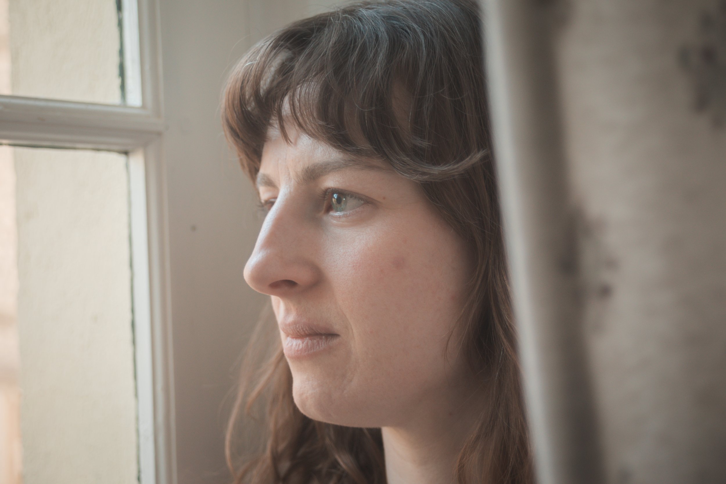 Une femme avec les cheveux bruns, regardant à travers une fenêtre, expression contemplative, intérieur, lumière naturelle.