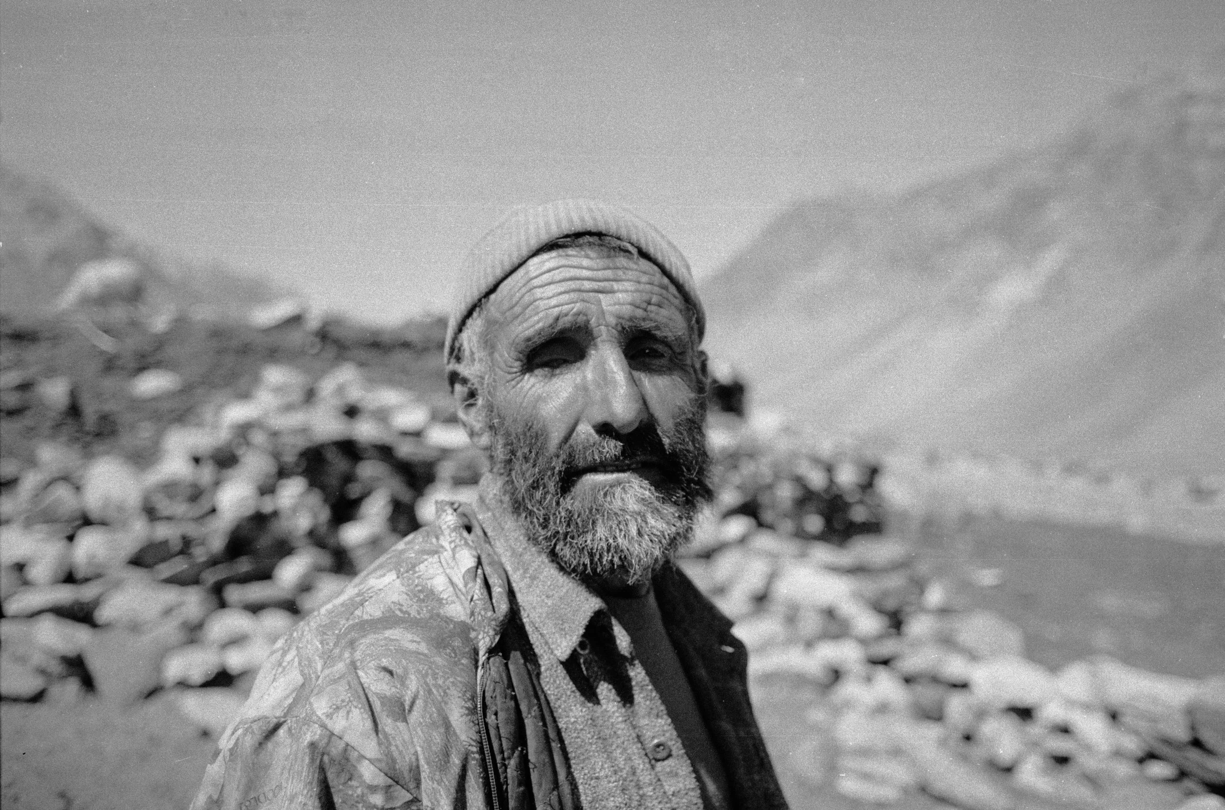 Portrait en noir et blanc d'un homme âgé avec une barbe, portant une casquette et un manteau, se tenant devant un paysage rocheux et un ciel nuageux.