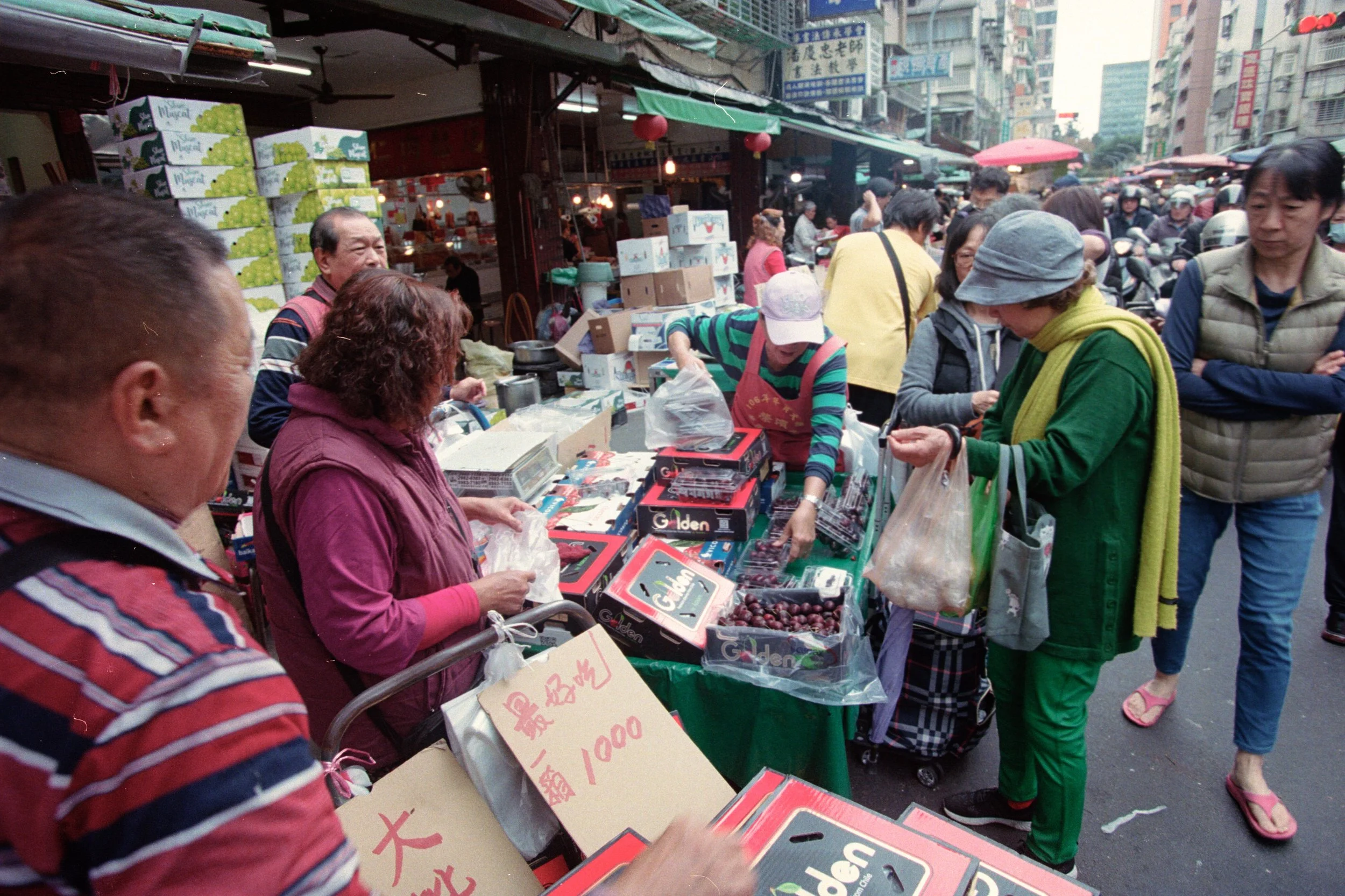 Marché de rue animé avec des étals vendant des fruits, des légumes et d'autres produits, entouré de nombreuses personnes faisant leurs achats dans un quartier urbain asiatique.