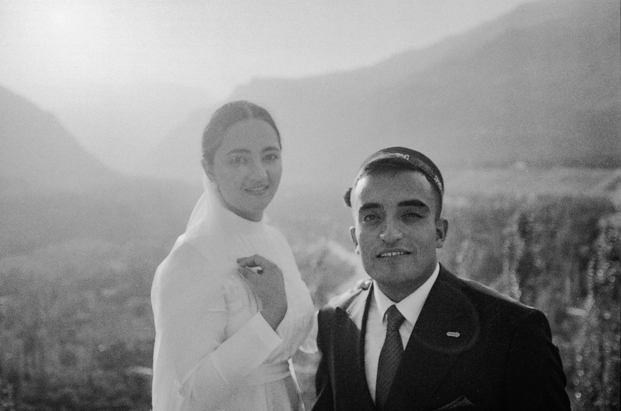 Deux personnes, une femme en robe blanche et un homme en costume noir, posent ensemble dans un paysage naturel.
