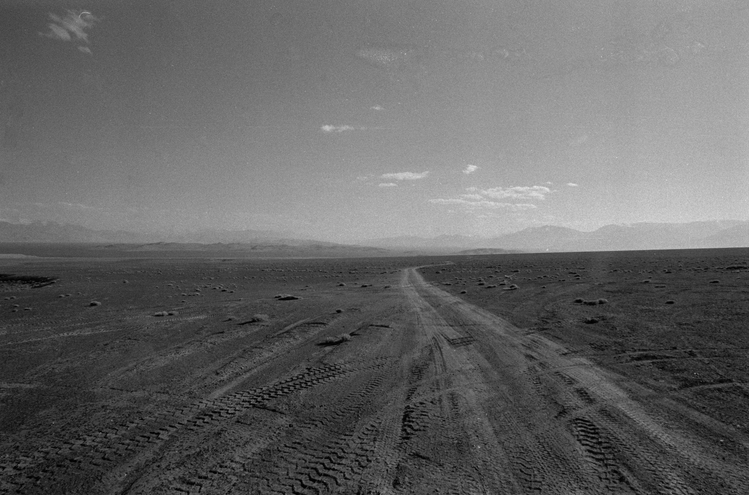 Un chemin dans un paysage désertique avec des montagnes au loin et un ciel clair avec quelques nuages.