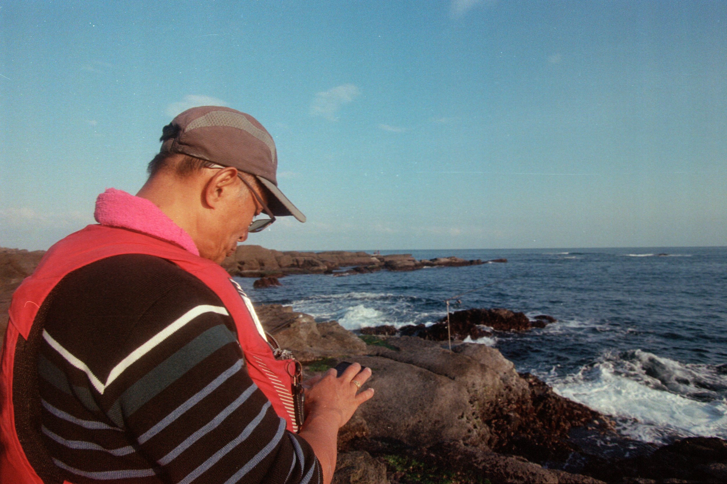 Une personne portant un maillot rayé, une veste rouge et une casquette grise regarde son téléphone sur un rivage rocheux à côté de la mer, ciel clair et horizon visible.