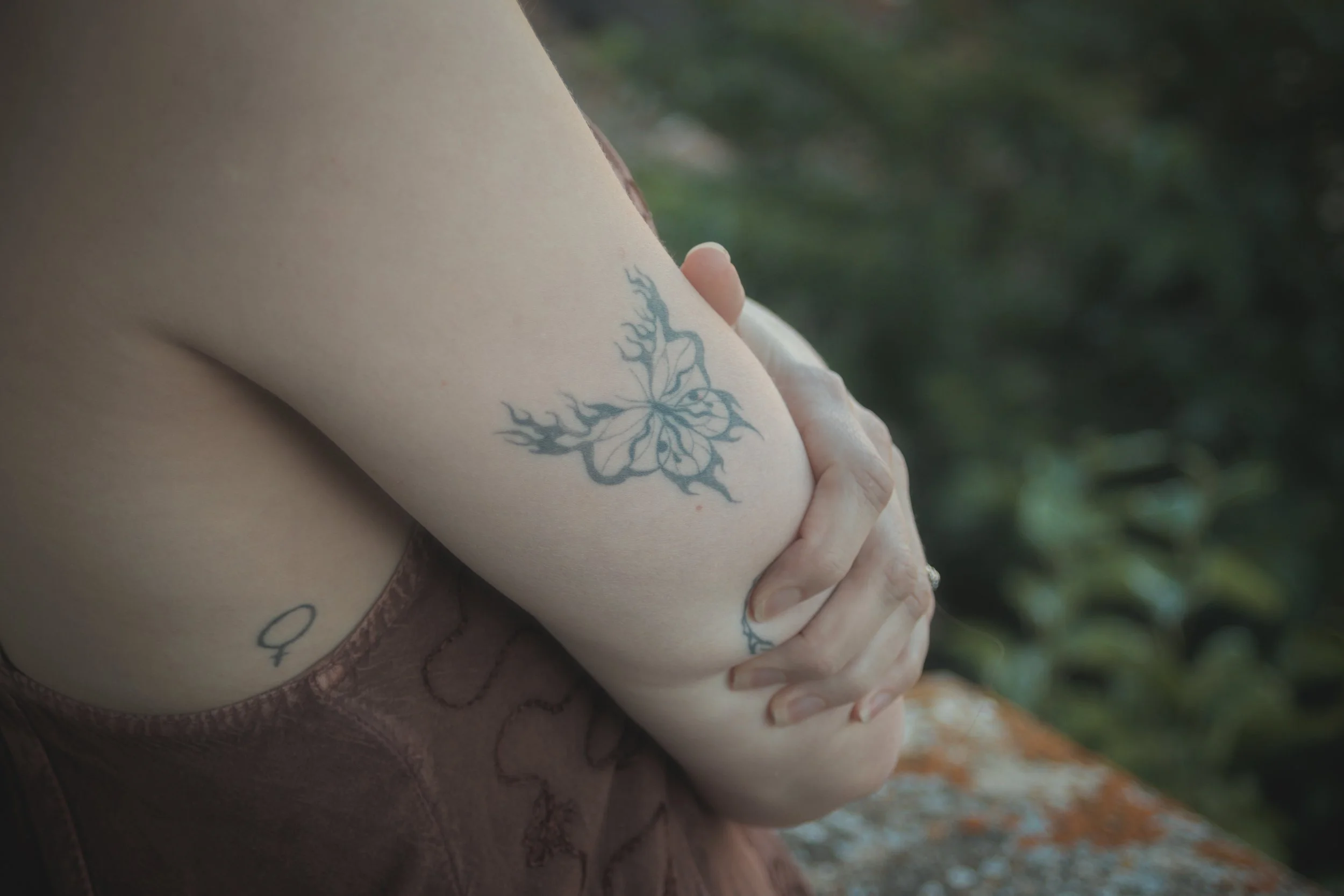 Un bras avec un tatouage d'une bisou dans un style délicat, tenant une autre personne dans un contexte extérieur avec un fond flou de nature.