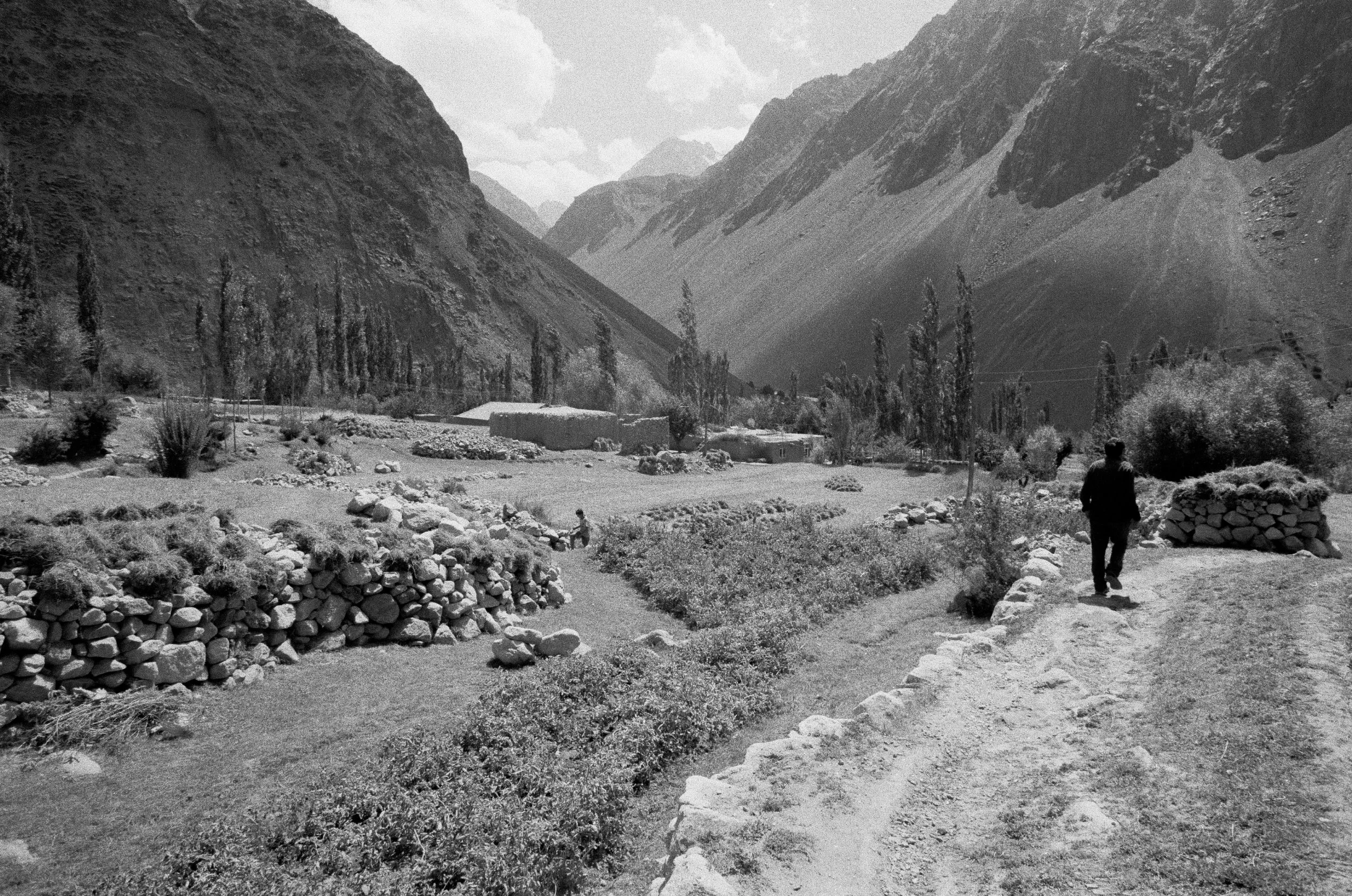 Vue d'une vallée montagneuse avec des maisons en bas, deux personnes marchant sur un sentier, et des montagnes escarpées en arrière-plan.