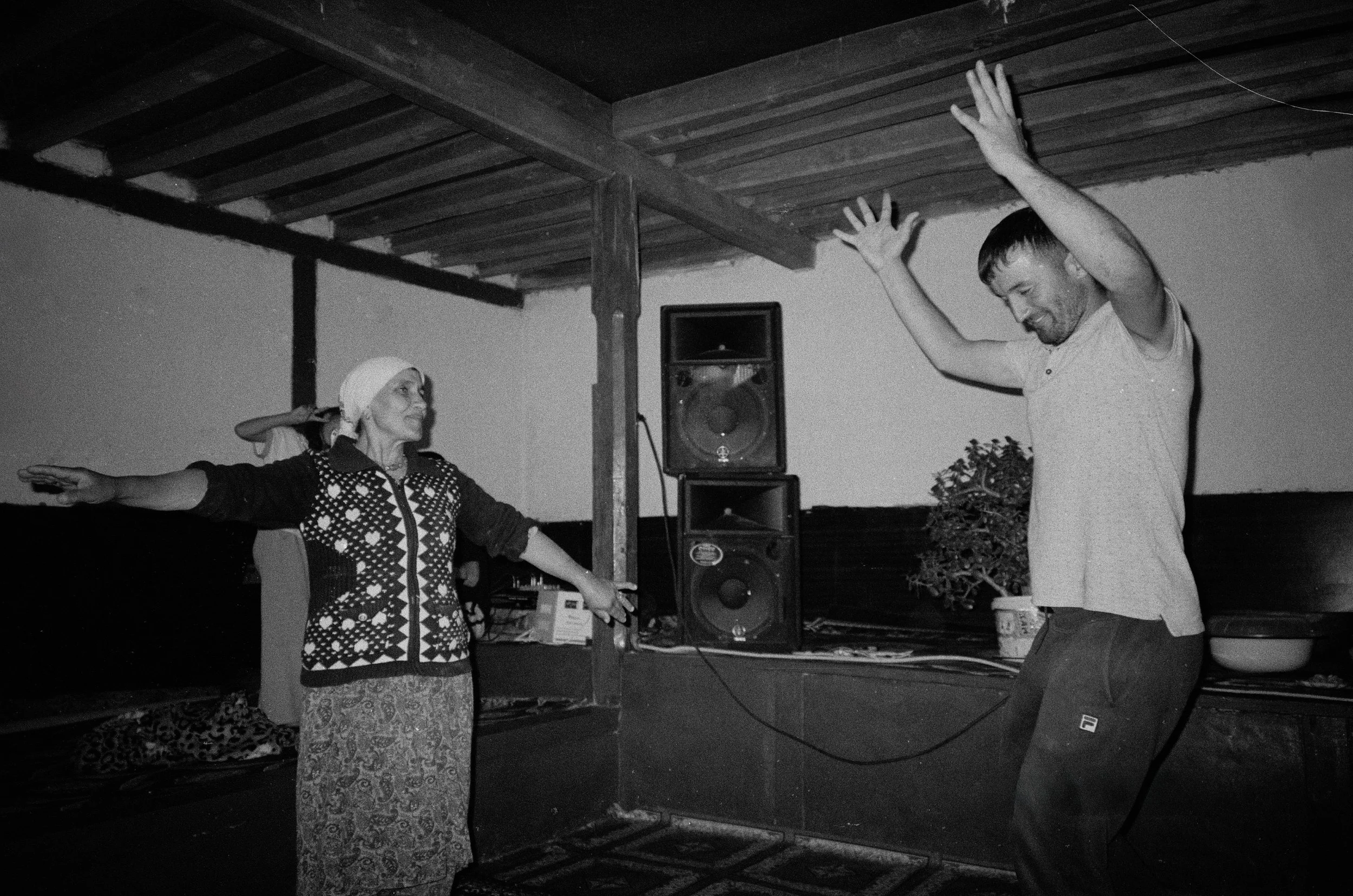 Un homme et une femme dansant dans une pièce avec un plafond en bois, une enceinte acoustique et un meuble avec des documents et des objets. La femme porte un pull à motifs et une coiffe blanche, l'homme porte un t-shirt blanc et des pantalons clairs
