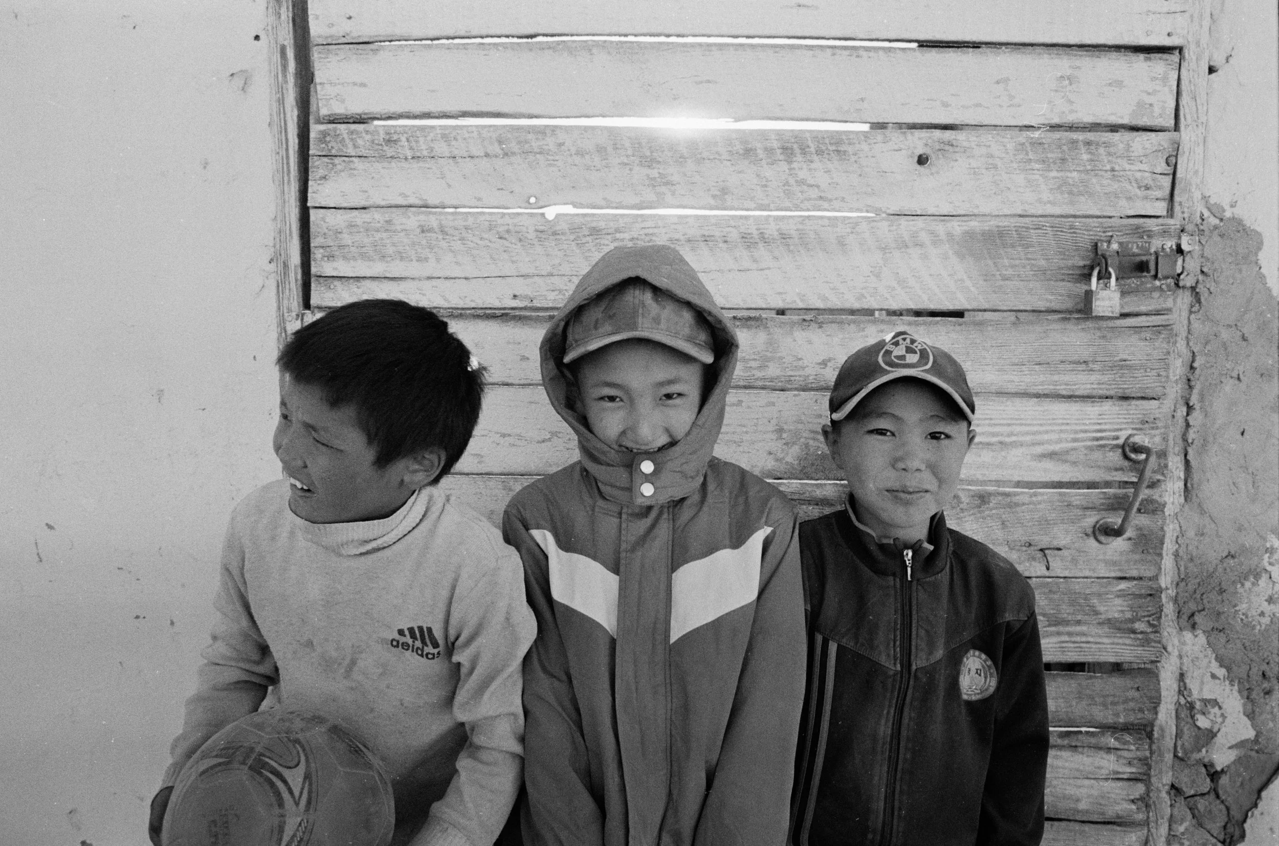 Quatre enfants souriants posent devant un mur en bois. Deux portent des casquettes, un porte une capuche, et l'autre regarde à gauche.