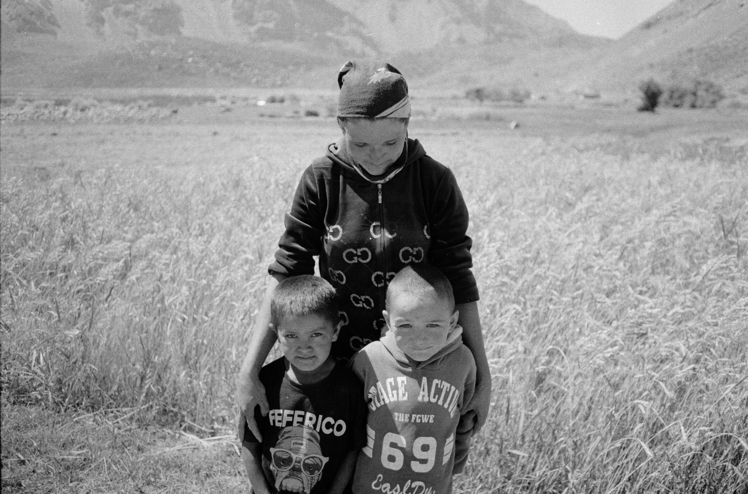Trois enfants jouent dans un champ d'herbe, deux sont à genoux et un est debout, tous regardant vers la caméra, avec des montagnes en arrière-plan.