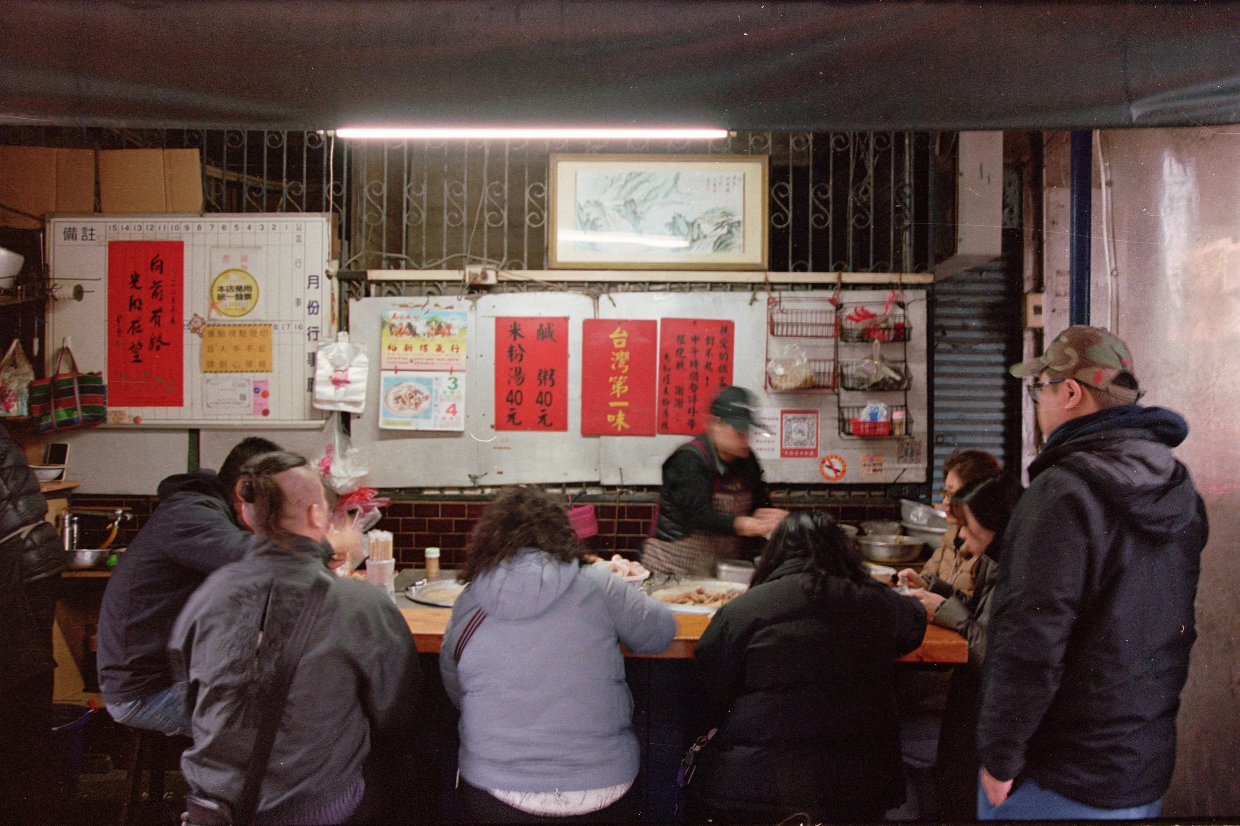 Un restaurant ou une salle à manger bondée avec des personnes assises autour de tables mangeant. Des affiches et des menus en chinois sont visibles sur le mur en arrière-plan.