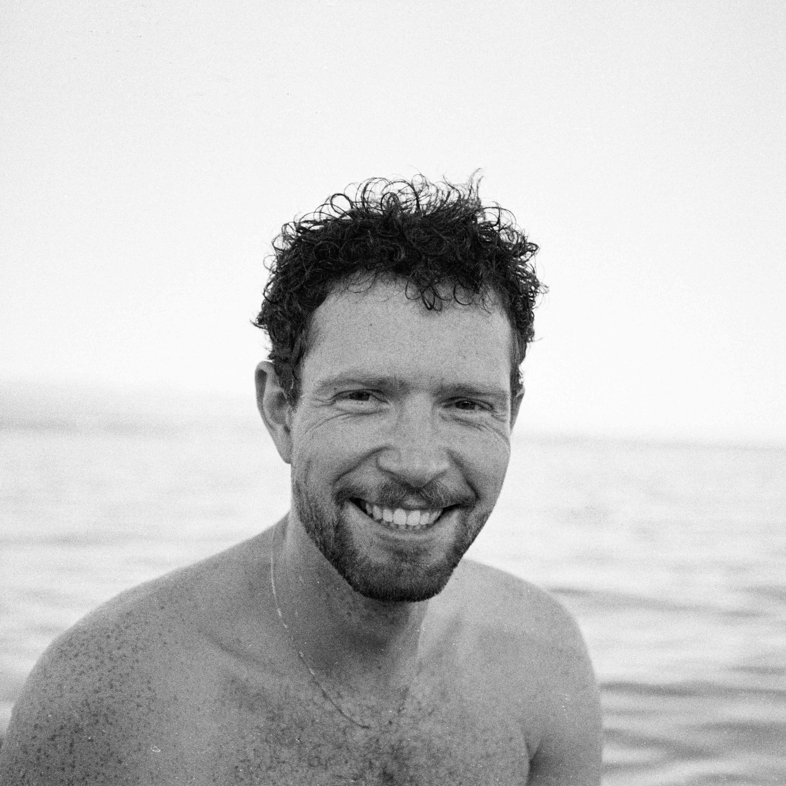 Un homme souriant avec une barbe, vu de près, portant un biker blanc, avec un fond blanc.