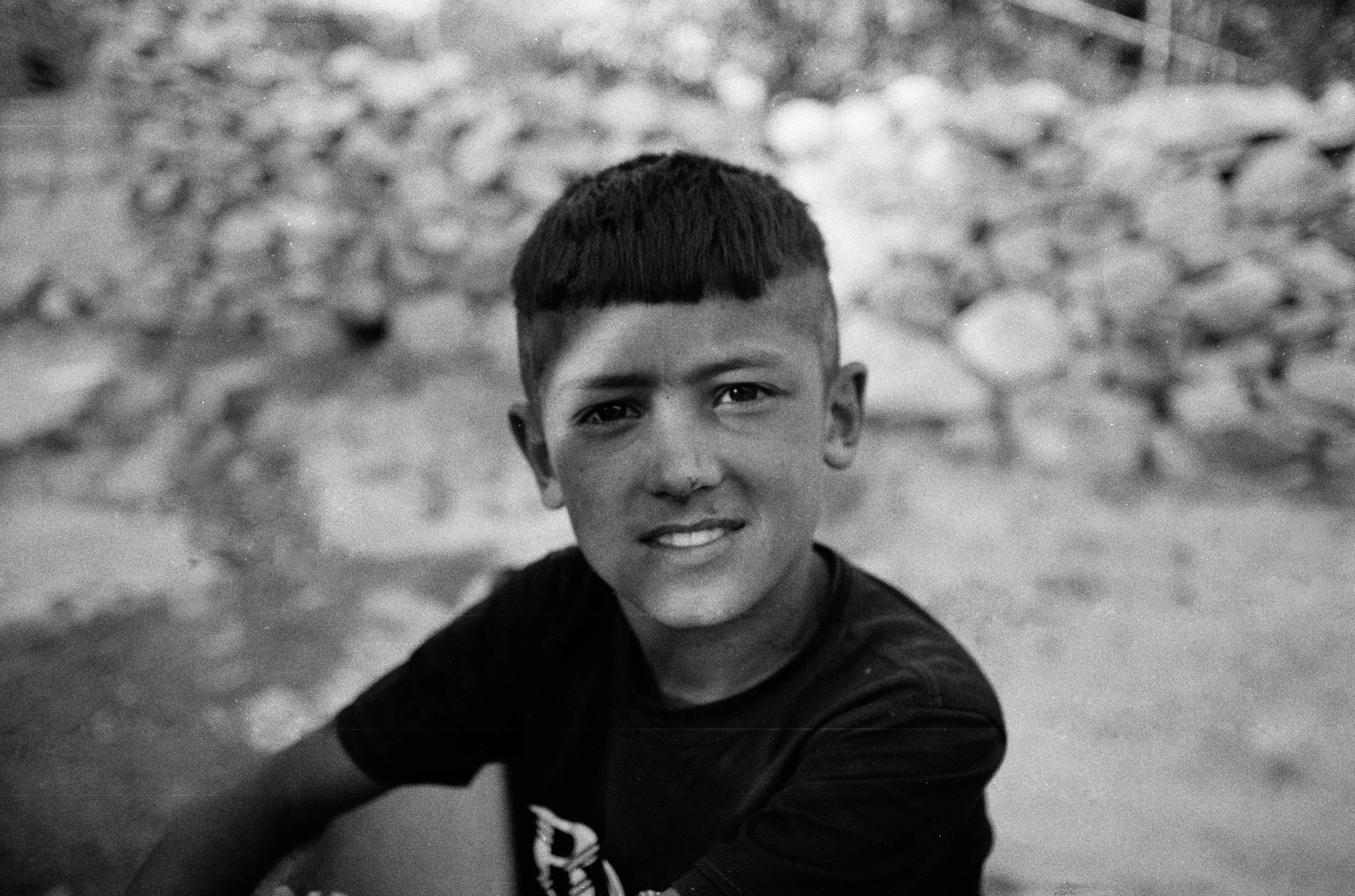 Portrait en noir et blanc d'un jeune garçon avec une expression souriante, portant un t-shirt noir, avec une étendue de galets ou de cailloux en arrière-plan.