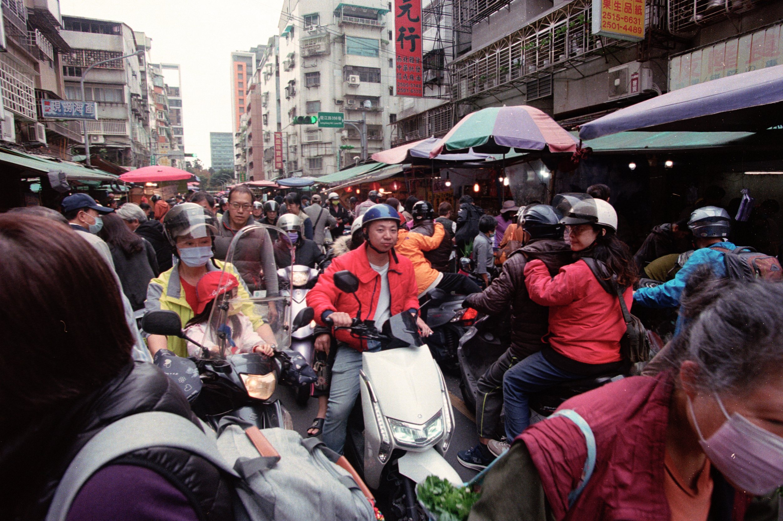 Foule de personnes portant des casques et des masques, en scooter ou à pied, dans une rue étroite et bondée d'une ville asiatique, avec des enseignes en écriture chinoise et des bâtiments variés