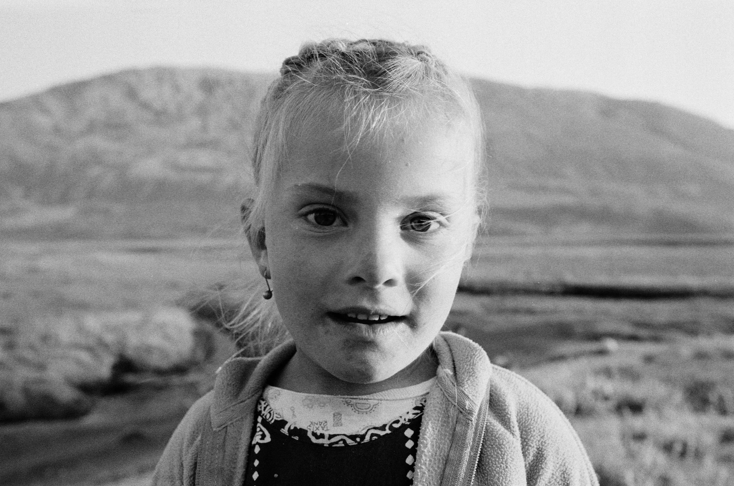 Un portrait en noir et blanc d'une jeune fille aux cheveux clairs, portant une veste et un foulard, avec un paysage de montagnes et de plage en arrière-plan.