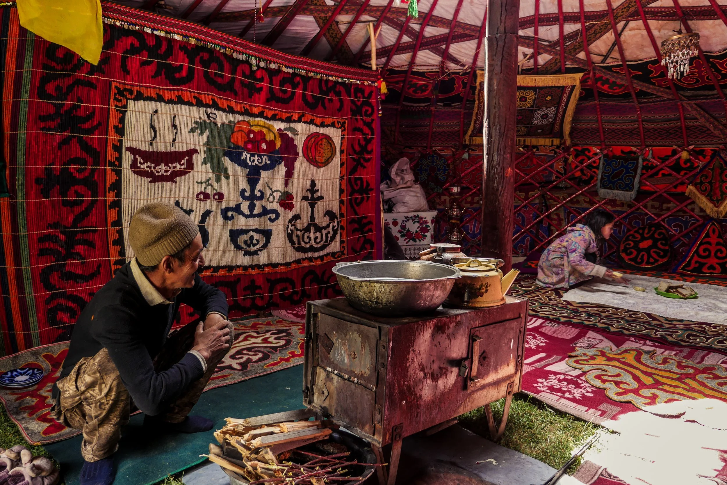 Une scène intérieure dans une tente traditionnelle, avec des tapis colorés aux motifs complexes accrochés aux murs et sur le sol. Un homme portant un bonnet est assis sur le tapis, peut-être en train de cuisiner ou de préparer quelque chose. À l'arrière, une jeune fille est assise, manipulant ou montrant un objet, avec diverses fournitures ou objets devant elle.