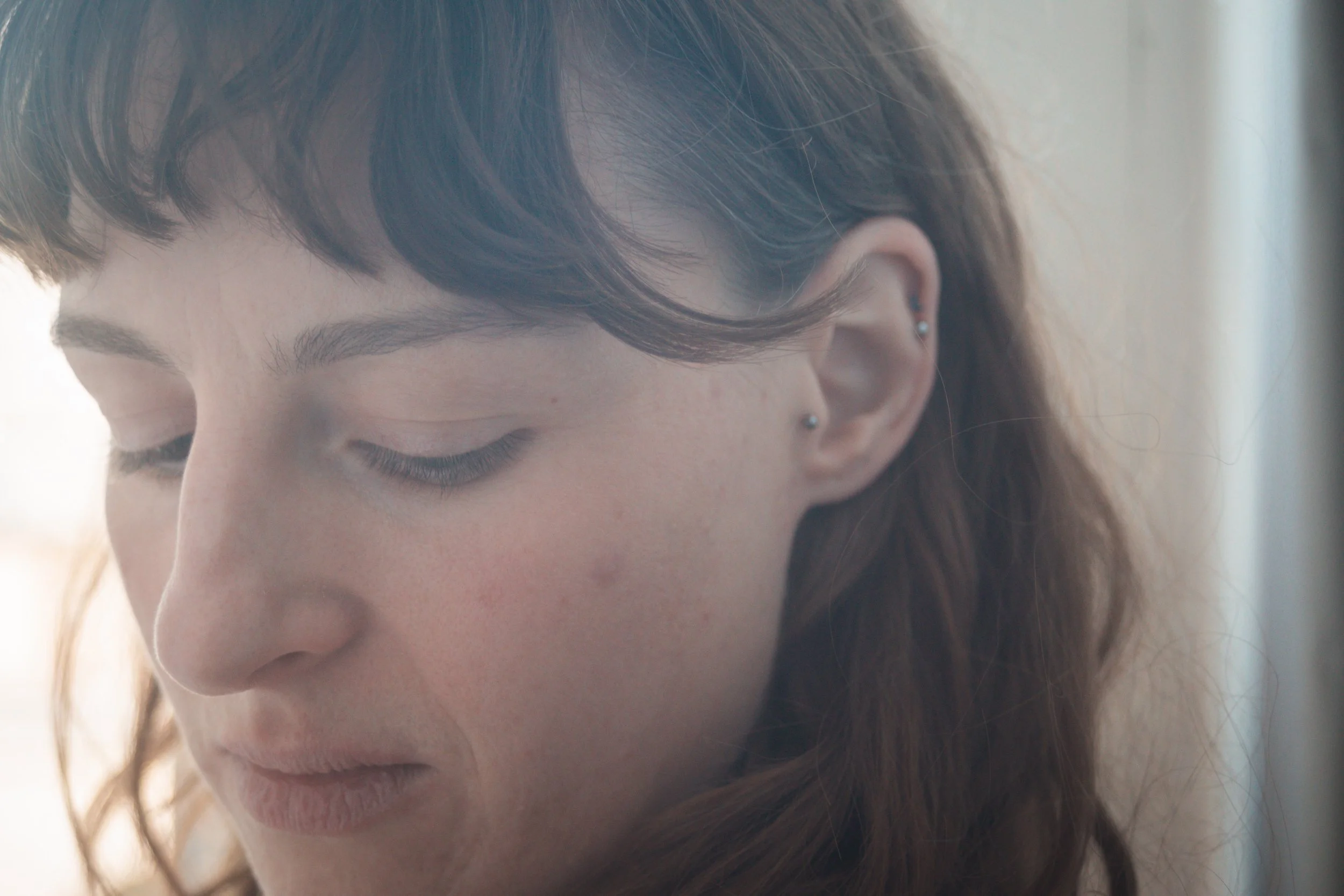 Close-up d'une femme avec les yeux fermés, cheveux bruns avec une boucle, portant une boucle d'oreille en perle, expression calme et pensive.