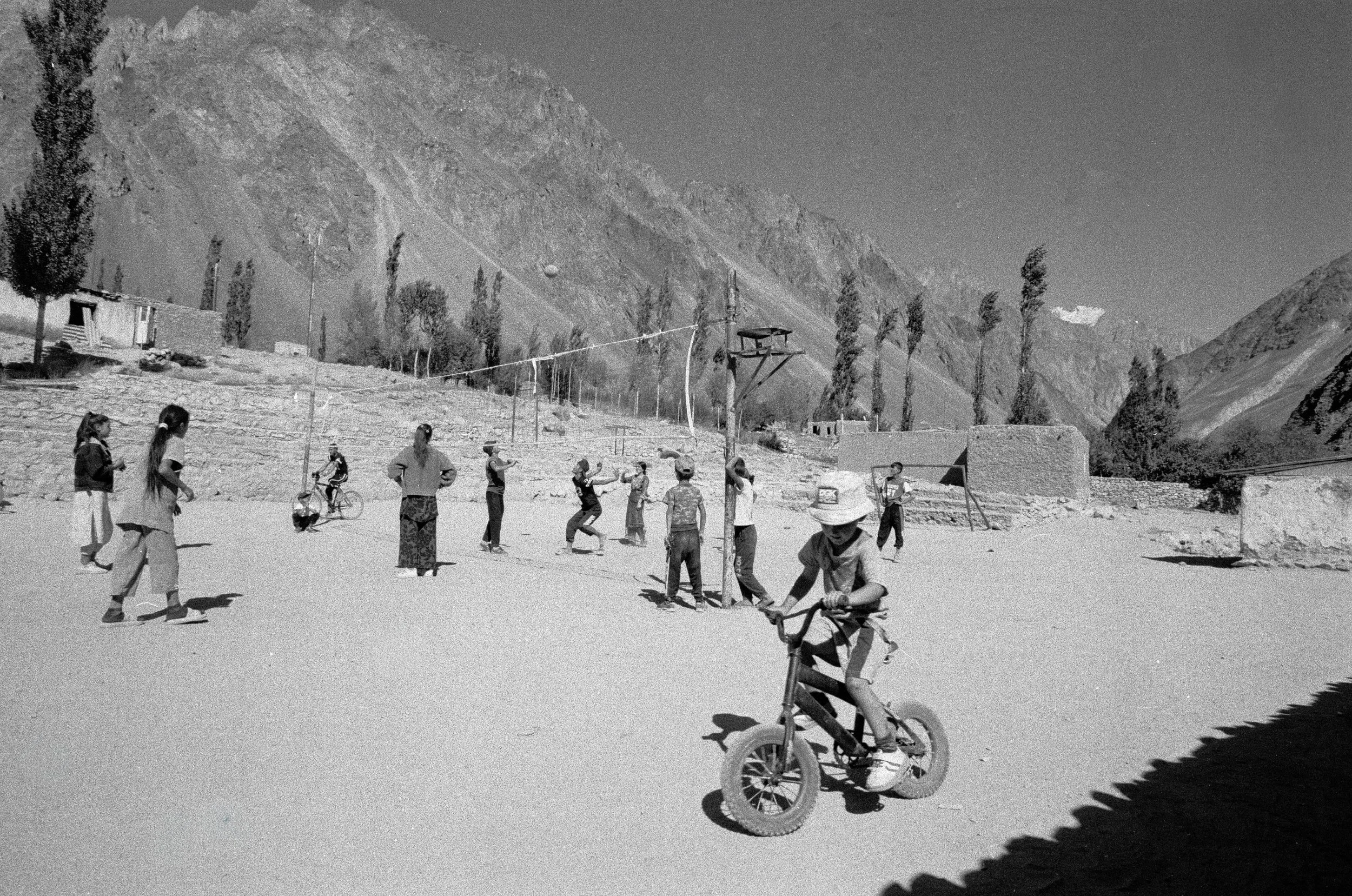 Des enfants et des adultes jouent et font du vélo dans un paysage montagneux, avec des montagnes en arrière-plan et des arbres.