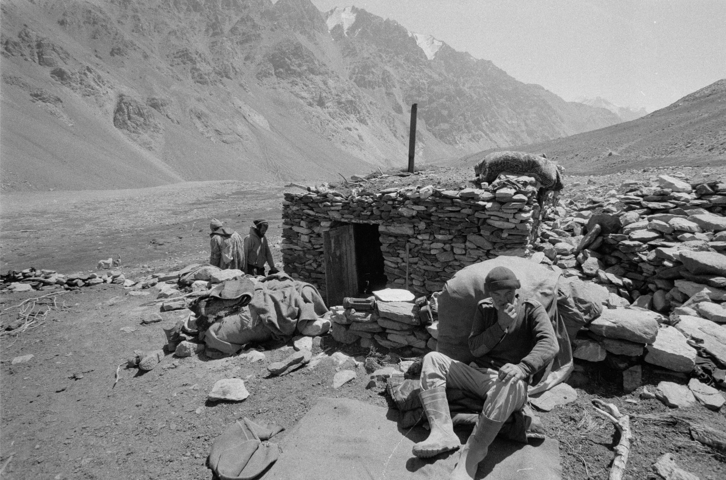 Paysage de montagne avec des rochers et des nuages, personnes en vêtements traditionnels assises et debout près d'une petite construction en pierre dans un environnement aride.