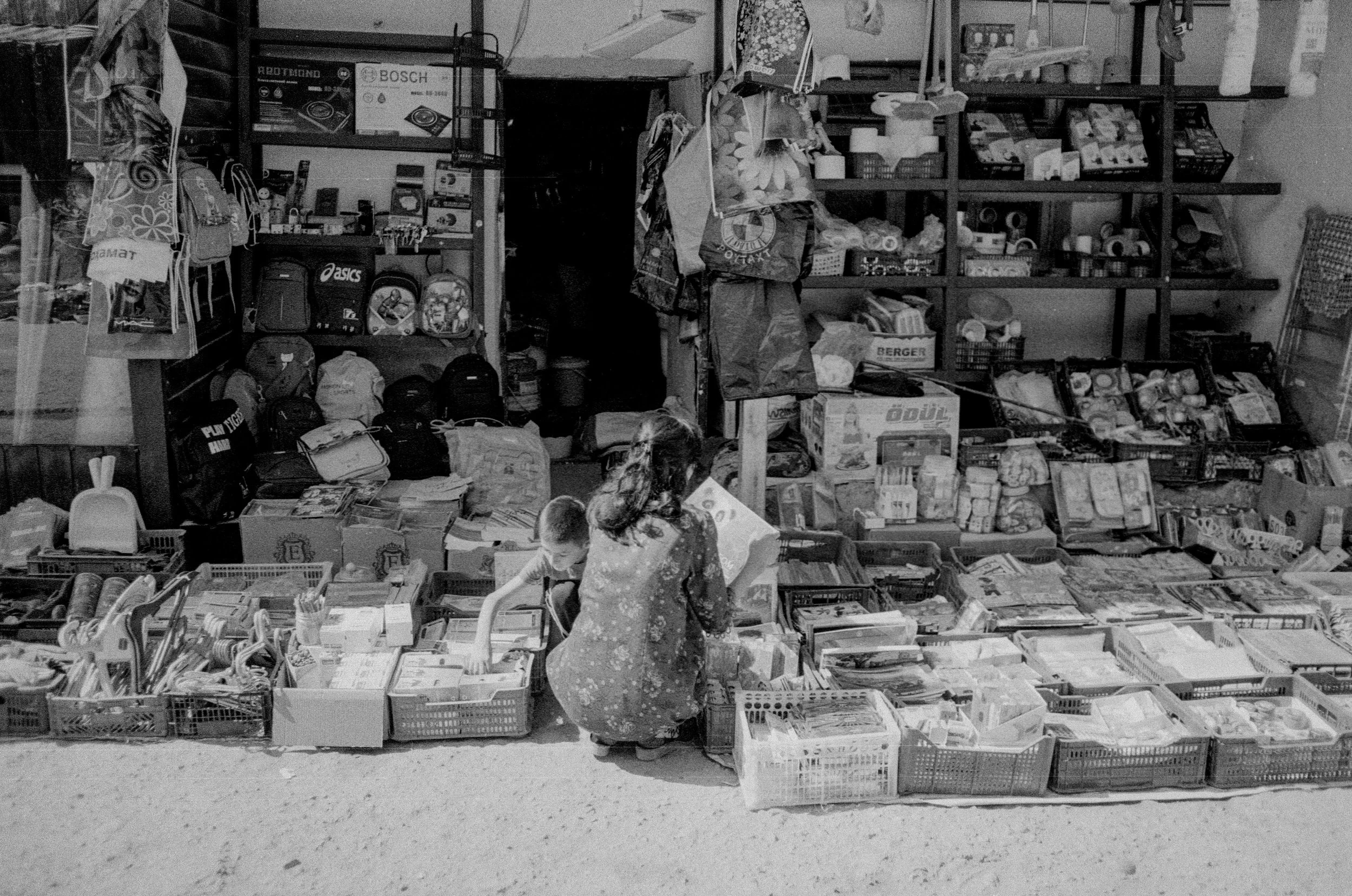 Une scène d'un magasin ou d'une librairie avec des étagères remplies de livres, sacs et autres articles. Deux enfants et une femme sont en train de regarder les articles sur une table.