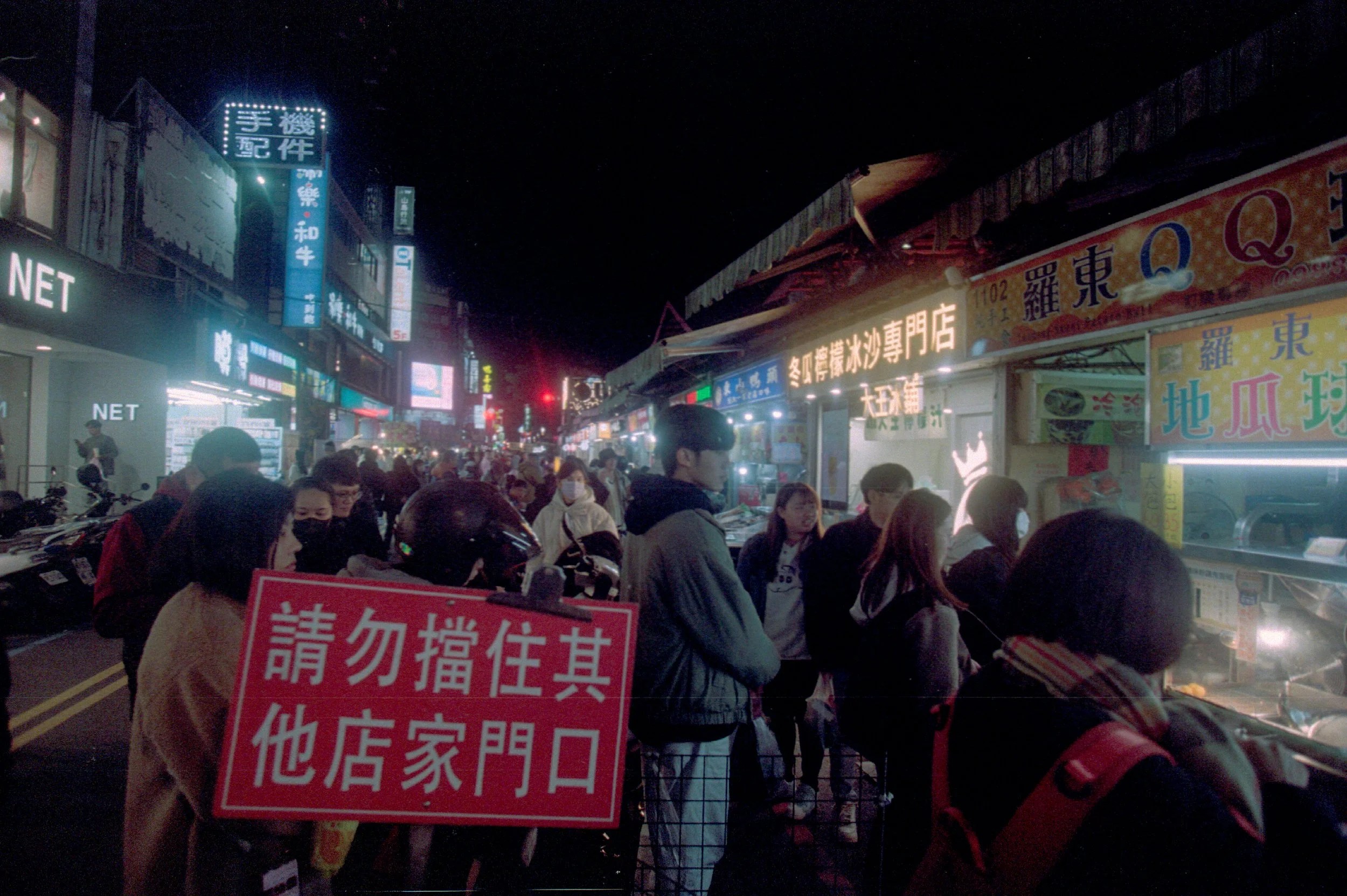 Une rue animée nocturne avec des enseignes lumineuses en chinois, de nombreuses personnes font la queue devant des stand de nourriture ou des magasins, certains portant des masques.