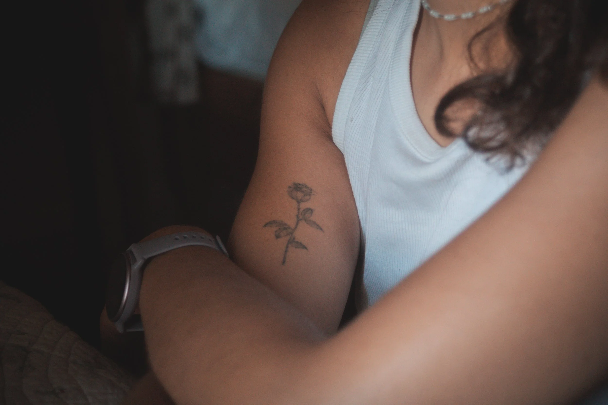 Vue rapprochée d'un bras d'une personne avec un tatouage de fleurs et d'insectes, portant un bracelet connecté, en posture inclinée ou en train d'écrire, portant un débardeur blanc, avec des cheveux bouclés.