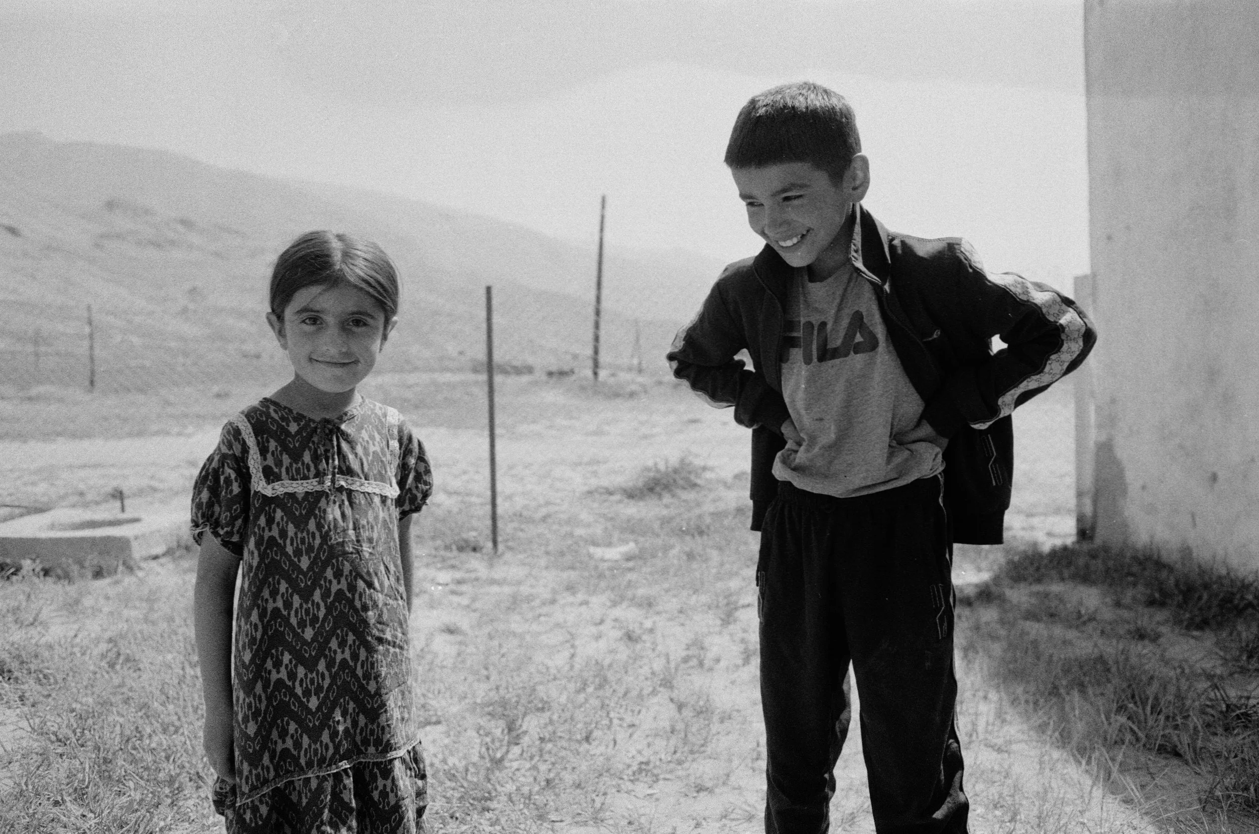 Deux enfants, une fille et un garçon, souriants, se tenant dans un champ outdoors, le garçon portant un sweat-shirt Fila et la fille portant une robe à motifs.