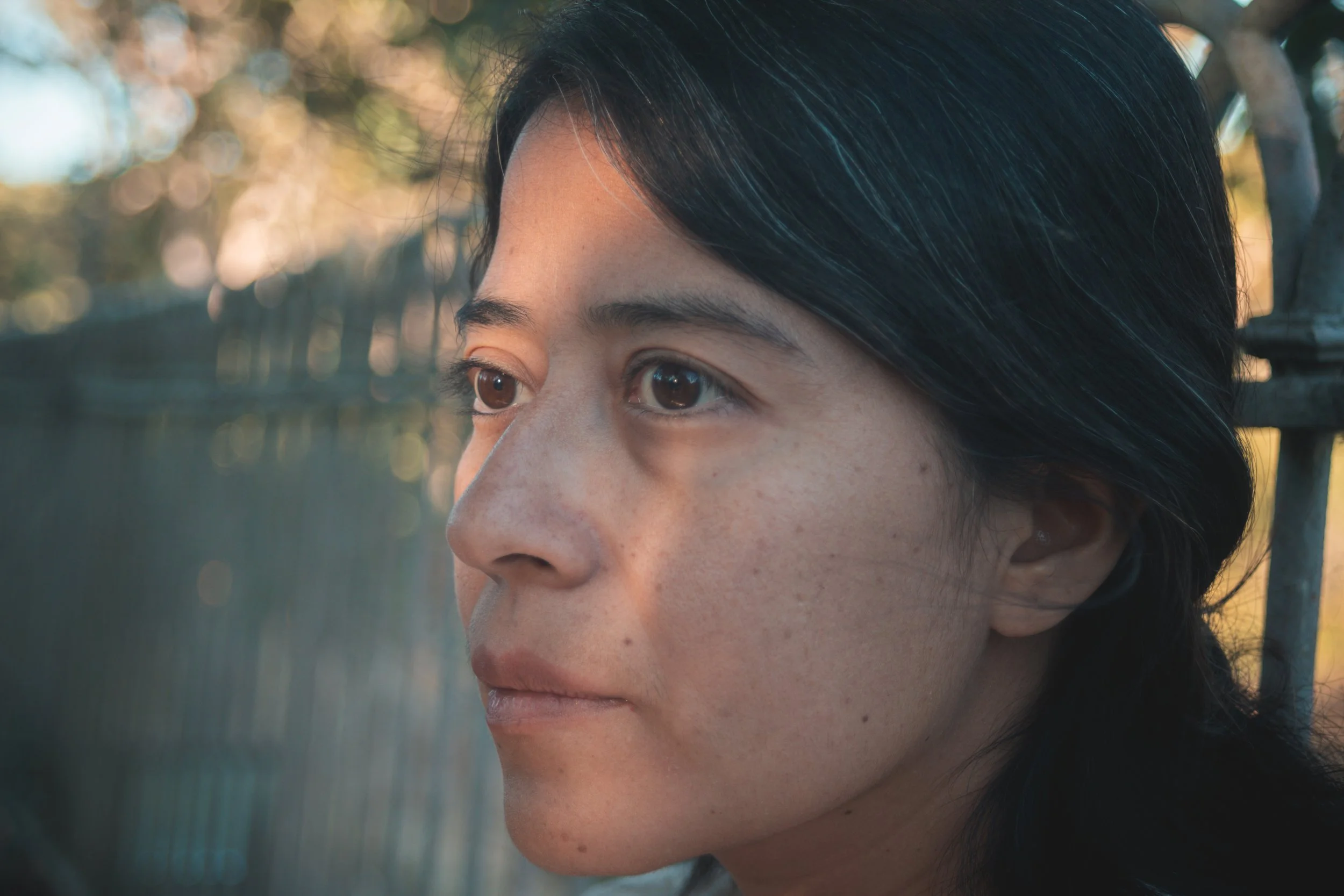 Portrait d'une femme asiatique regardant vers la gauche, avec un fond flou d'arbres et de lumière naturelle.