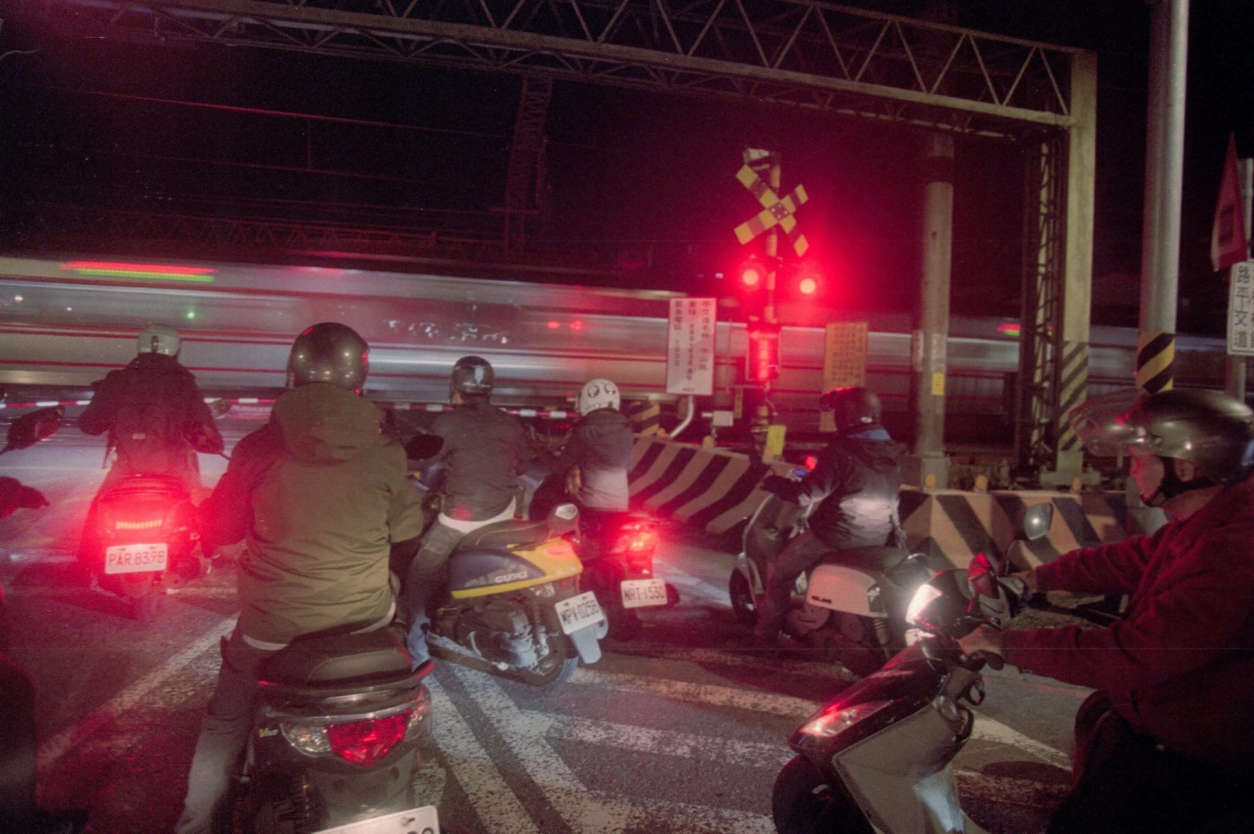 Groupe de personnes à moto arrêtées au niveau d'une barrière de train avec signal lumineux rouge dans la nuit.