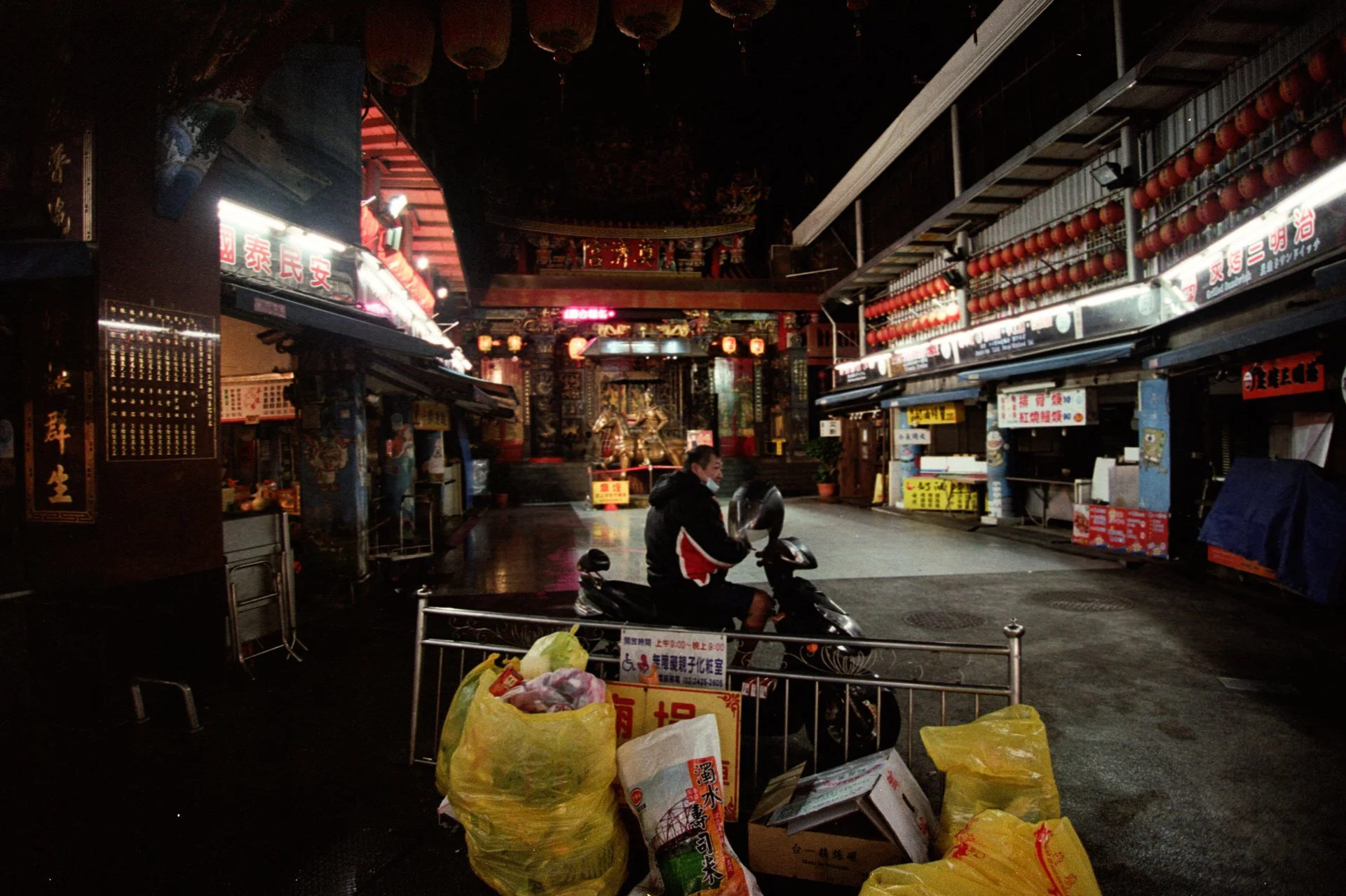 Une rue commerçante la nuit avec des lanternes rouges suspendues, un homme sur une moto devant des stands fermés, des sacs jaunes en premier plan, enseignes en caractères chinois, un intérieur décoré dans un style traditionnel chinois.