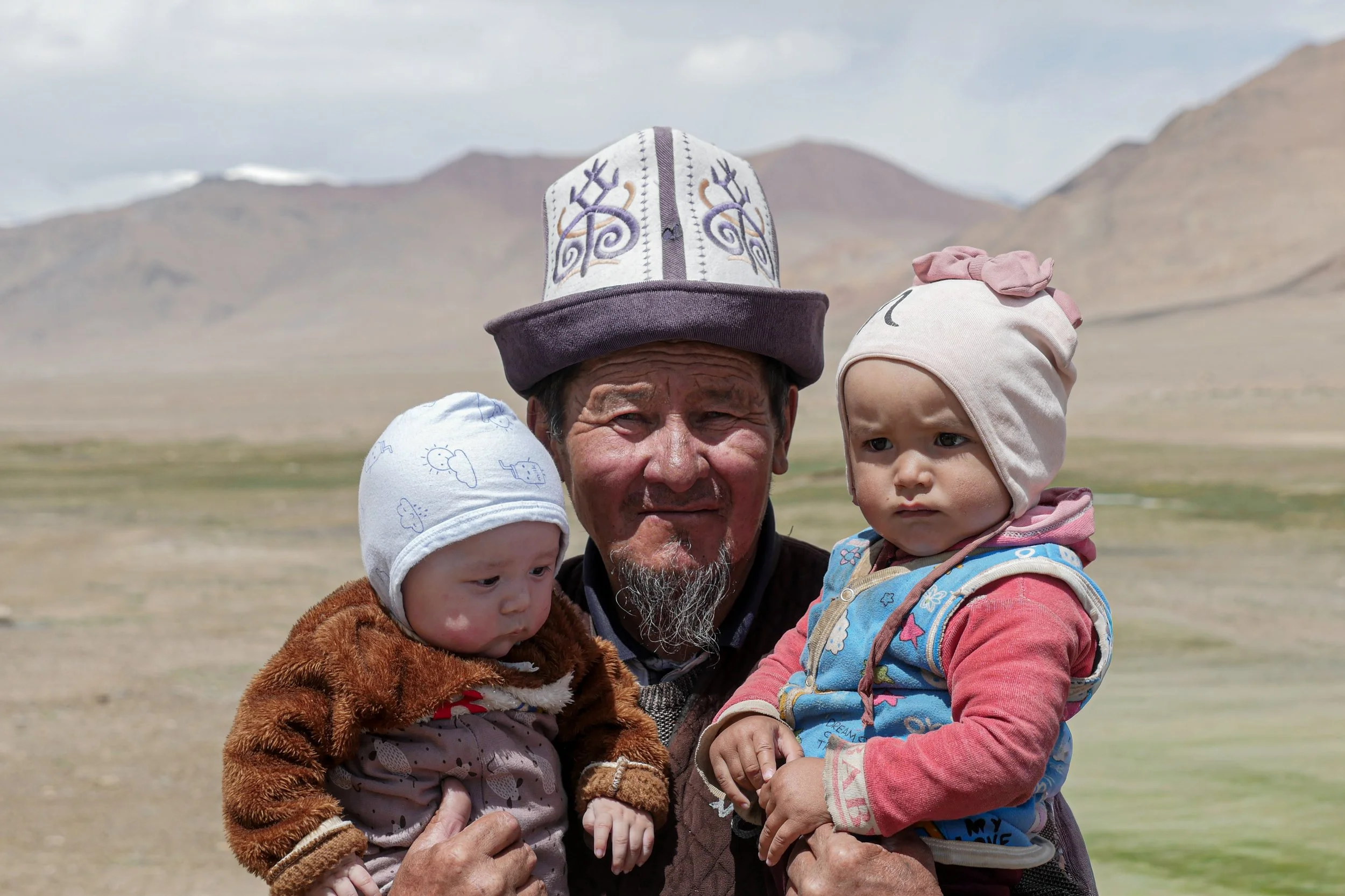 Un homme avec deux bébés, l'un dans chaque bras, en milieu rural avec des montagnes en arrière-plan.