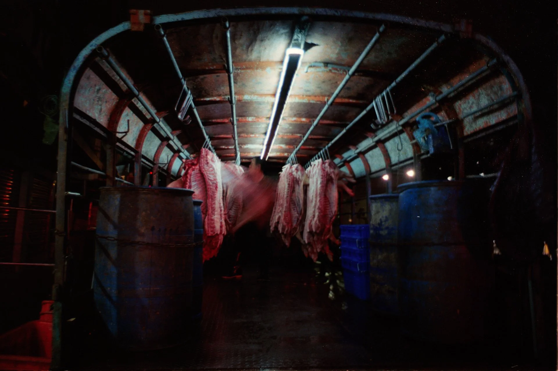Tournée de viande suspendue dans une chambre froide avec des bidons en métal et des caisses en plastique bleues.