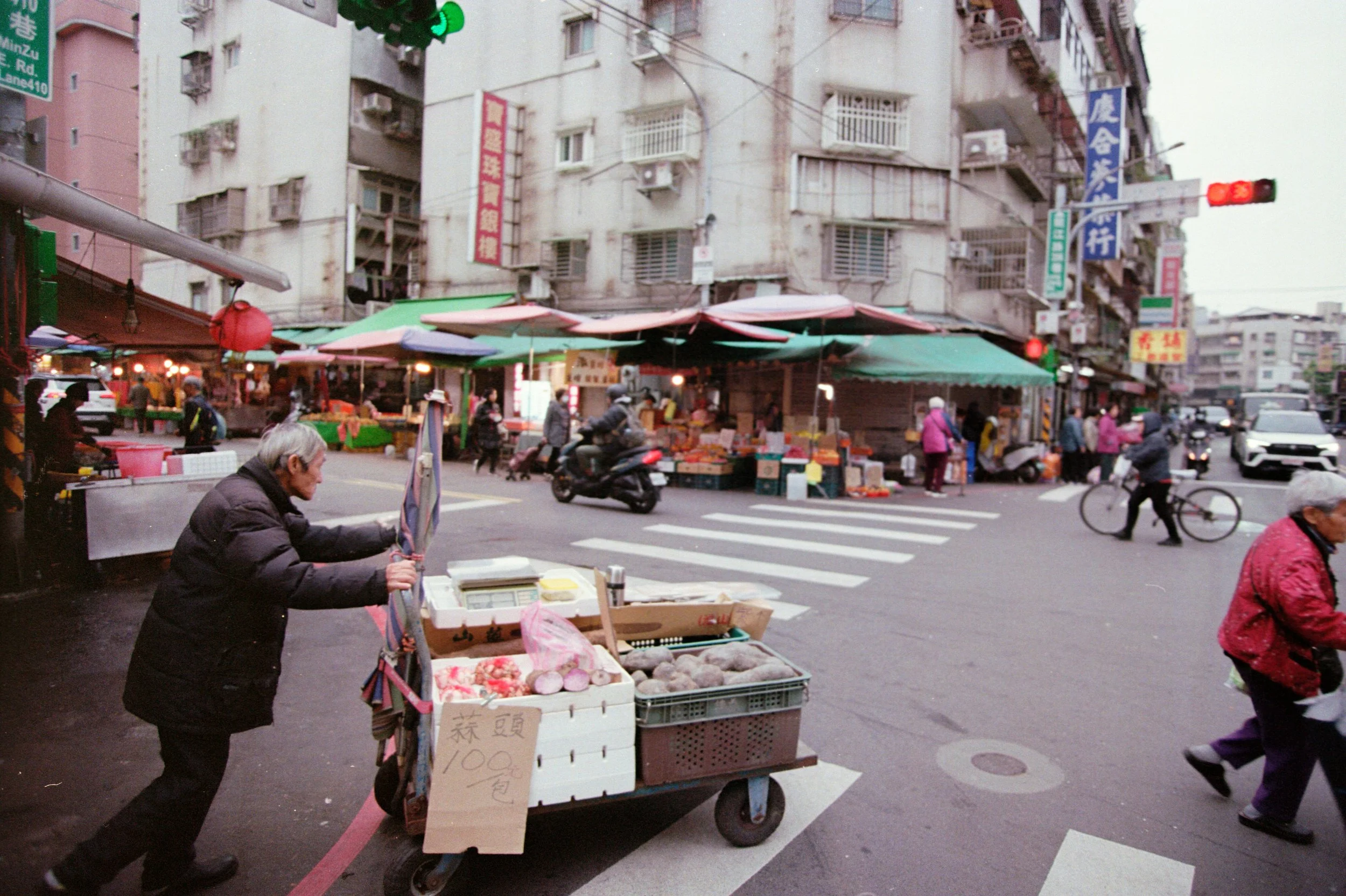 Un marché en plein air avec des étals de fruits et légumes, des vendeurs et des passants dans une rue urbaine.