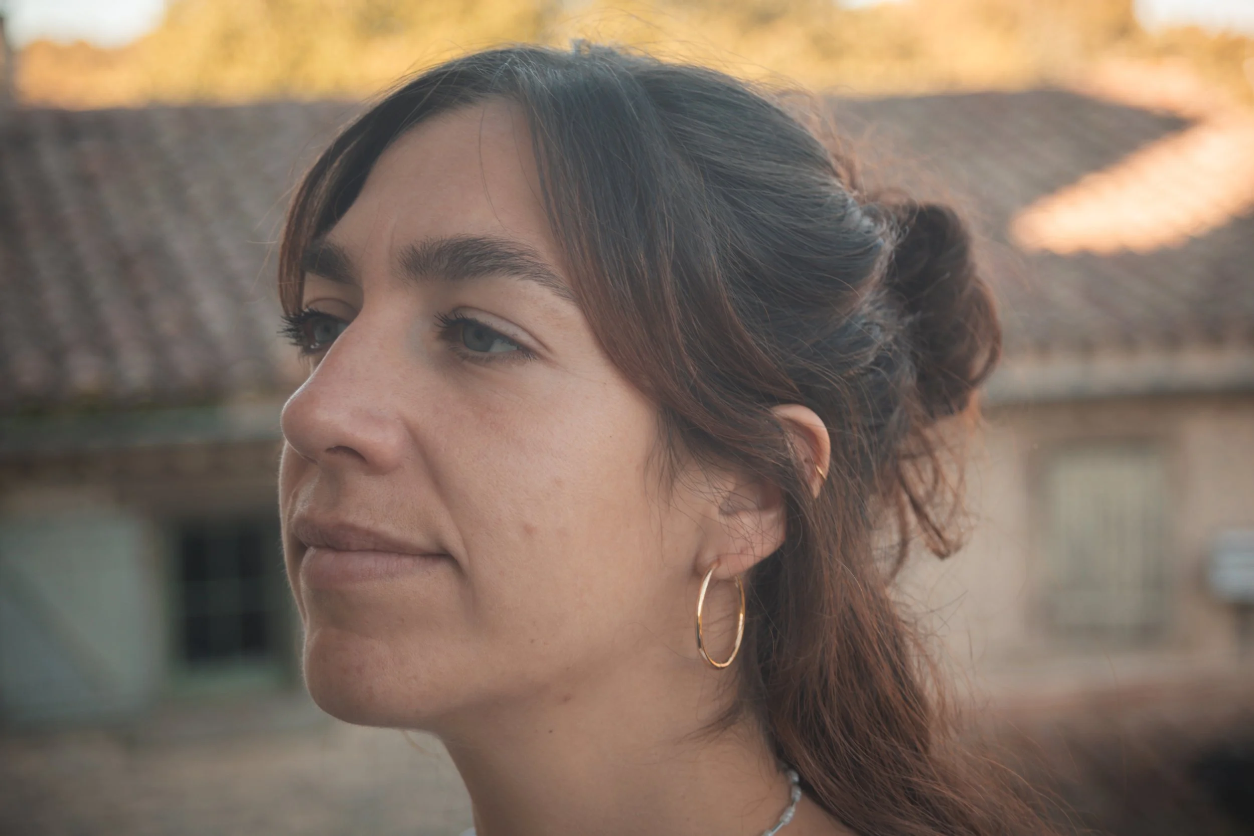 Portrair d'une femme aux cheveux bruns bouclés, portant une boucle d'oreille en or et un collier, avec un fond flou de nature en automne.