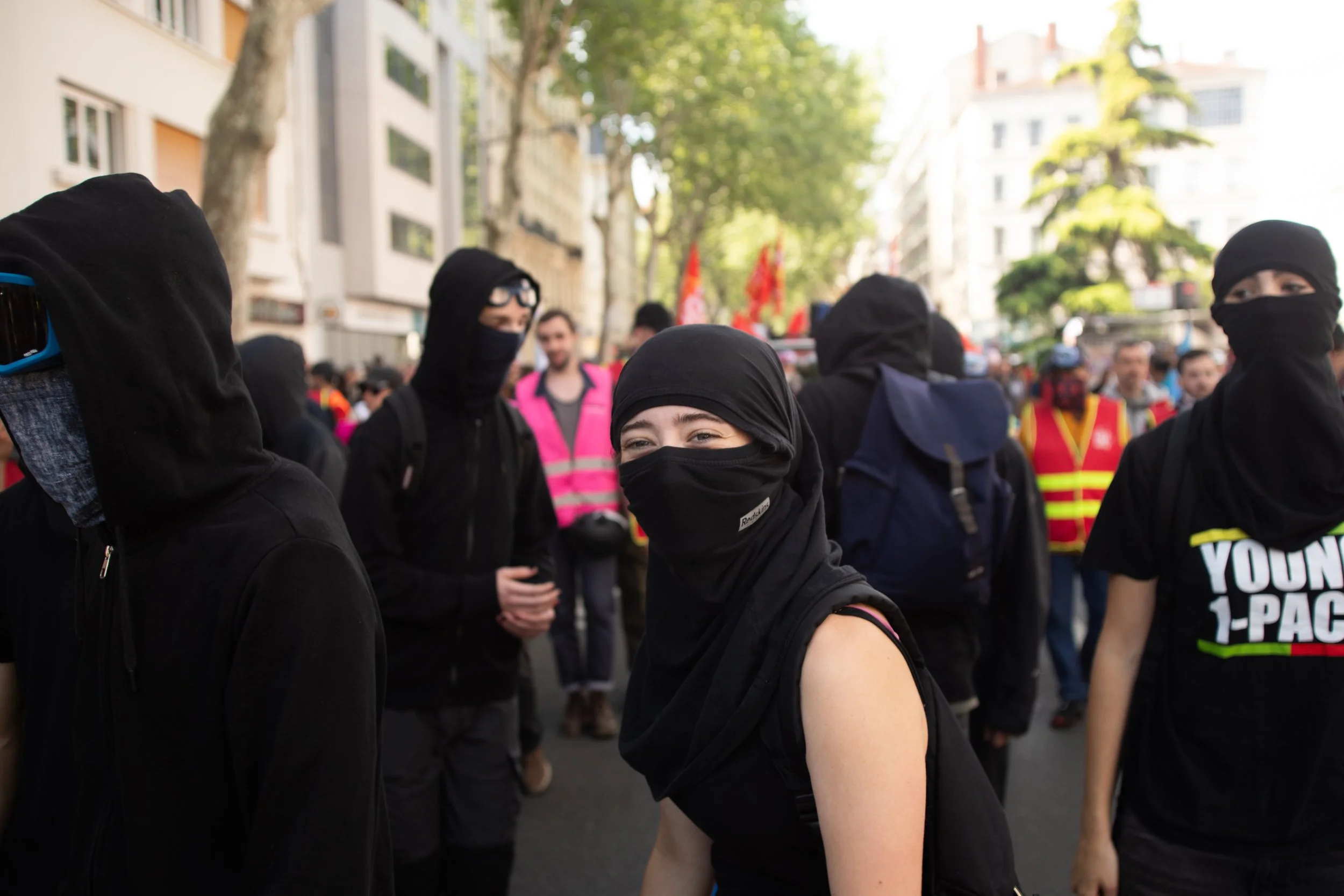 Des manifestants portant des masques noirs dans une manifestation urbaine, avec des bâtiments et des arbres en arrière-plan.