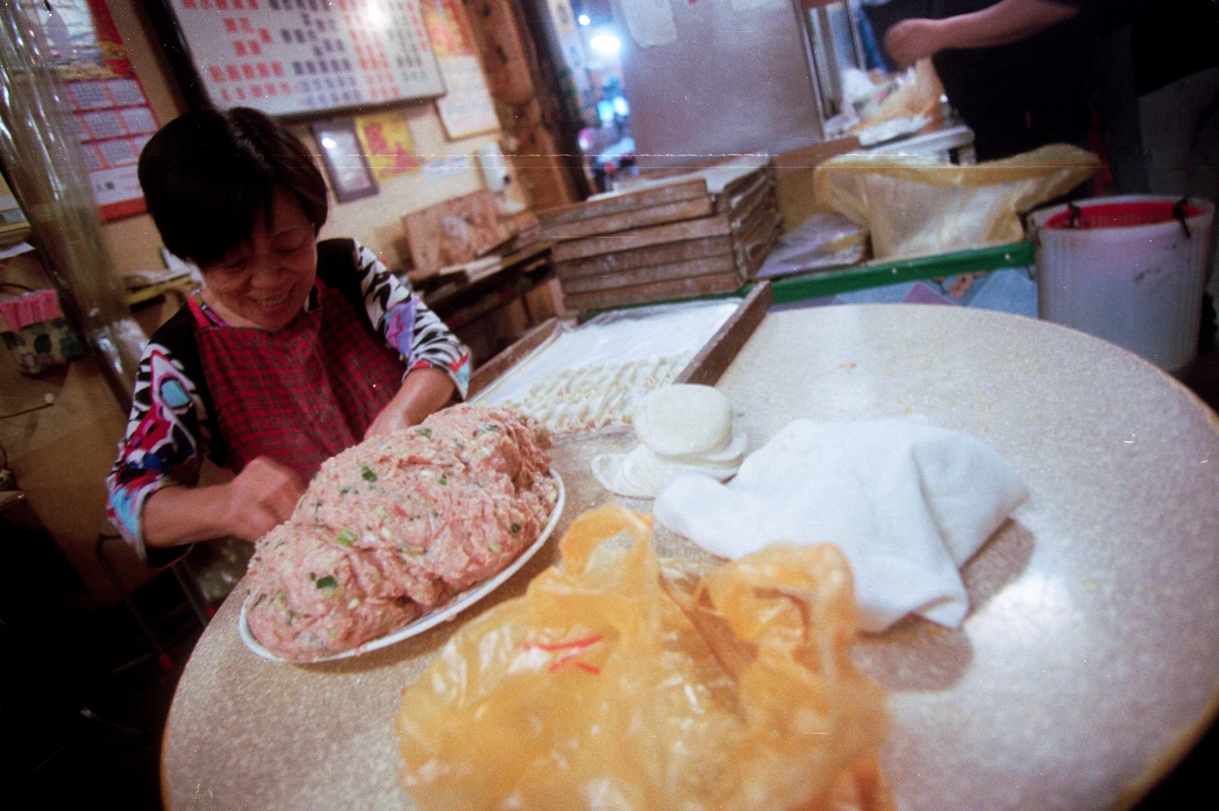 Une femme en train de préparer une pâte ou un mélange dans une assiette, dans une cuisine ou un espace de préparation alimentaire, avec des sacs de farine ou de pâte sur le comptoir et divers ustensiles ou ingrédients en arrière-plan.