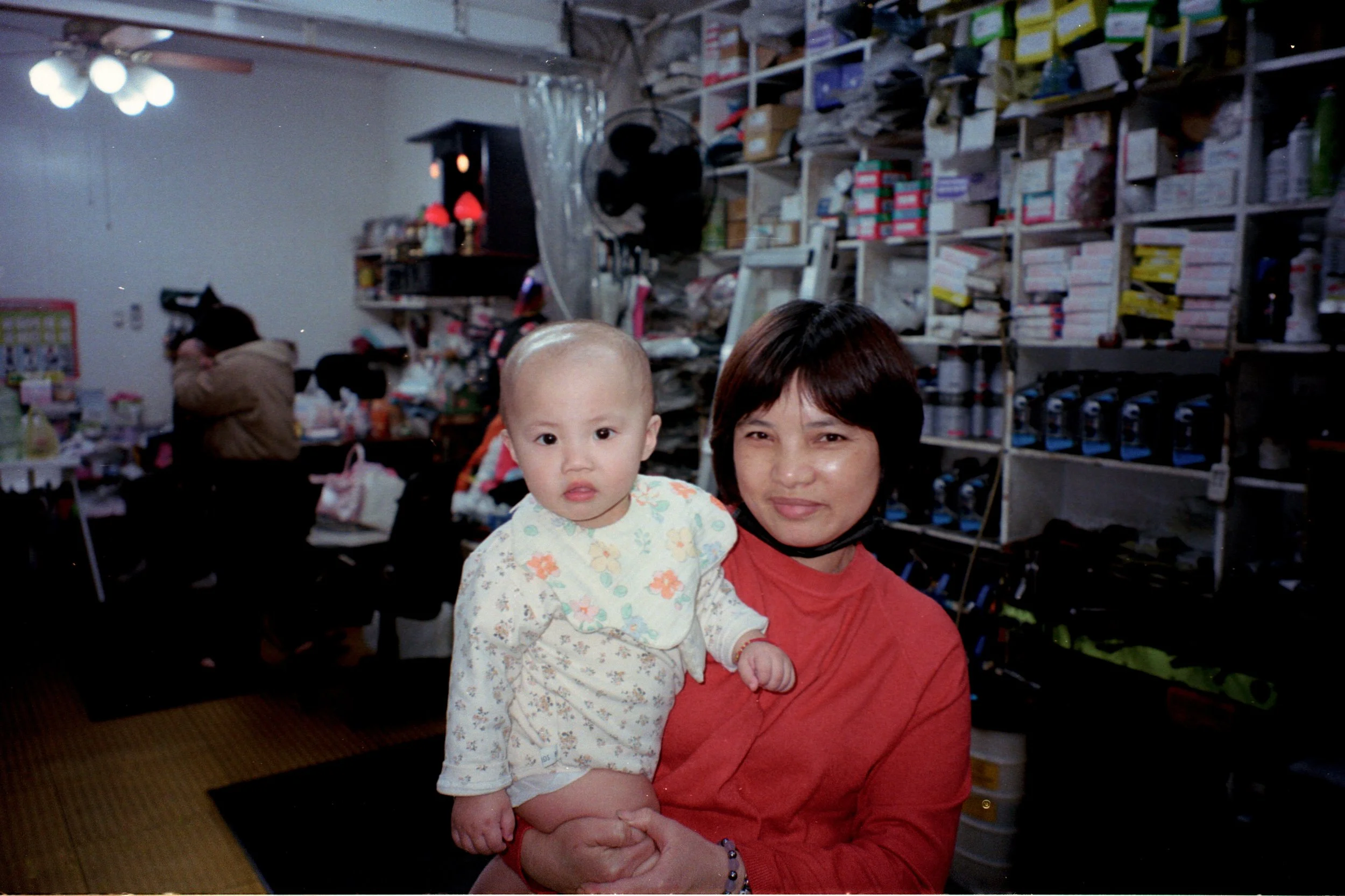 Une femme tient un jeune enfant dans une boutique ou un magasin rempli de rayonnages contenant divers produits. La femme sourit tandis que l'enfant regarde devant lui.
