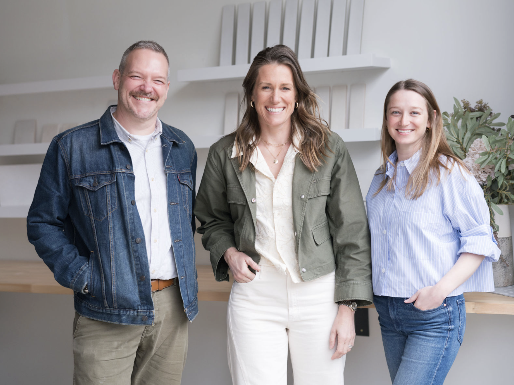 Three smiling people standing inside a modern, minimalist room with white walls, wooden surfaces, white shelves, and a plant in the background.