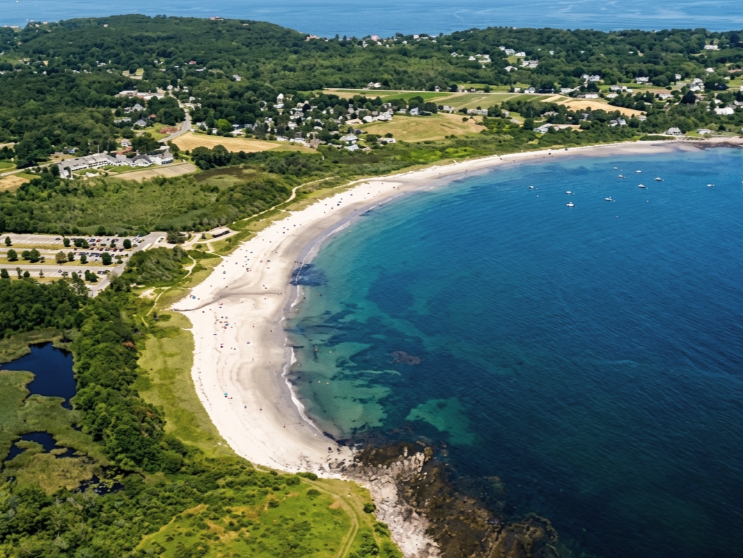 Aerial view of a crescent-shaped beach with white sand, surrounded by lush green trees and residential areas, along a calm blue ocean.